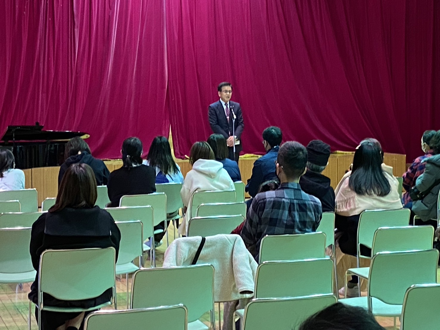 赤い幕を背景にスーツ姿の市長が壇上で話し、椅子に座った参加者たちが正面から耳を傾けている室内集会の写真