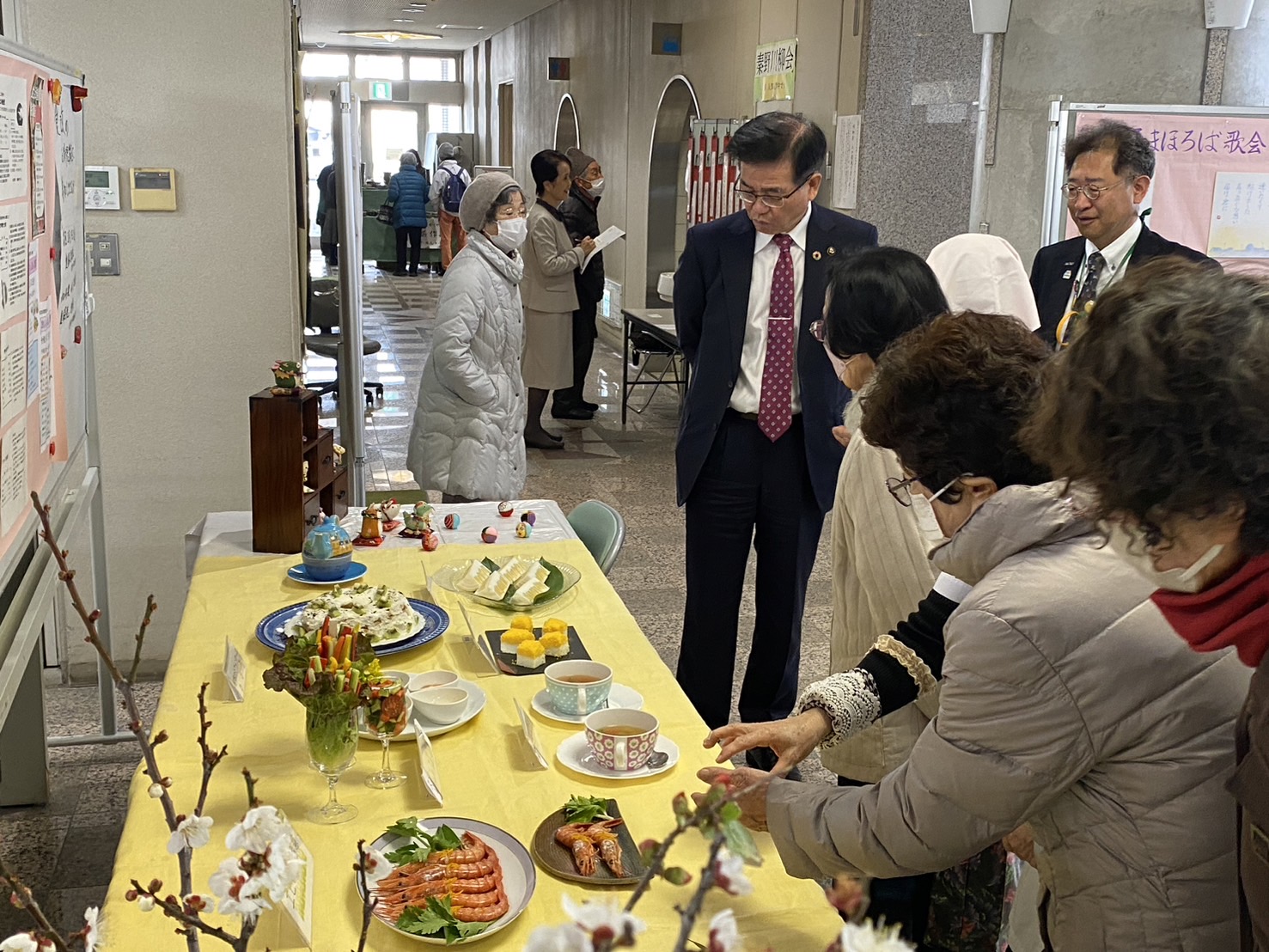 黄色いクロスの上に料理やお菓子が並ぶ展示台を人々が囲み、スーツ姿の市長も興味深そうに見学している室内イベントの写真