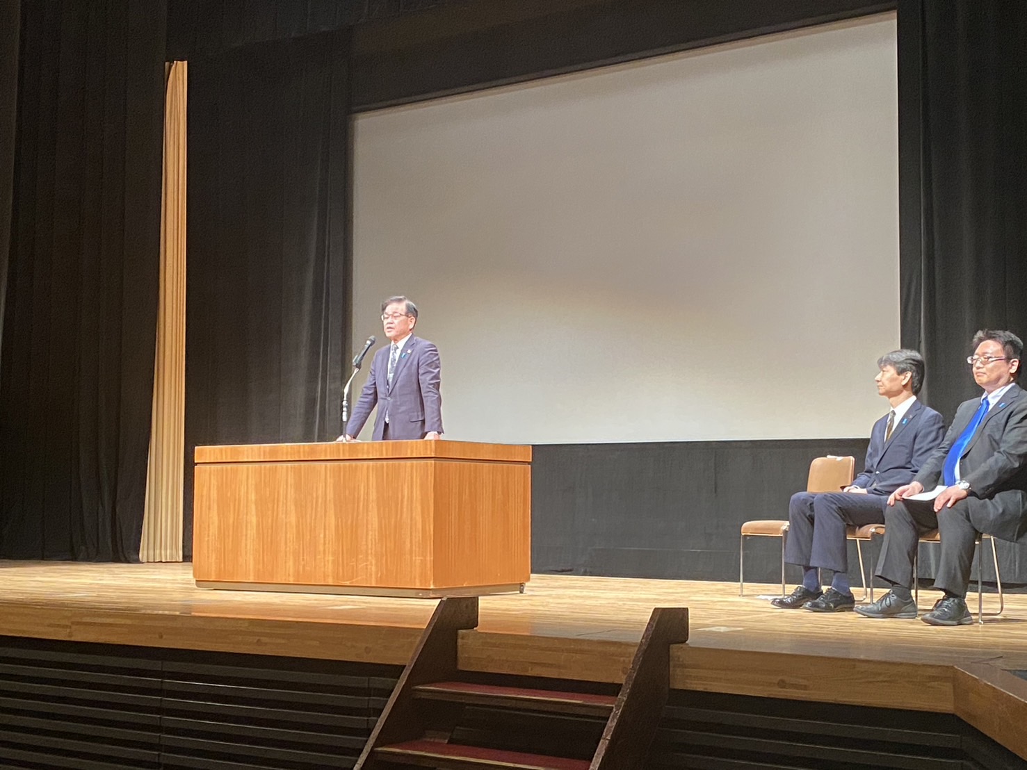 大きなスクリーンを背に演壇でスーツ姿の市長が挨拶し、横に座る関係者数名が見守るホールでの式典の写真