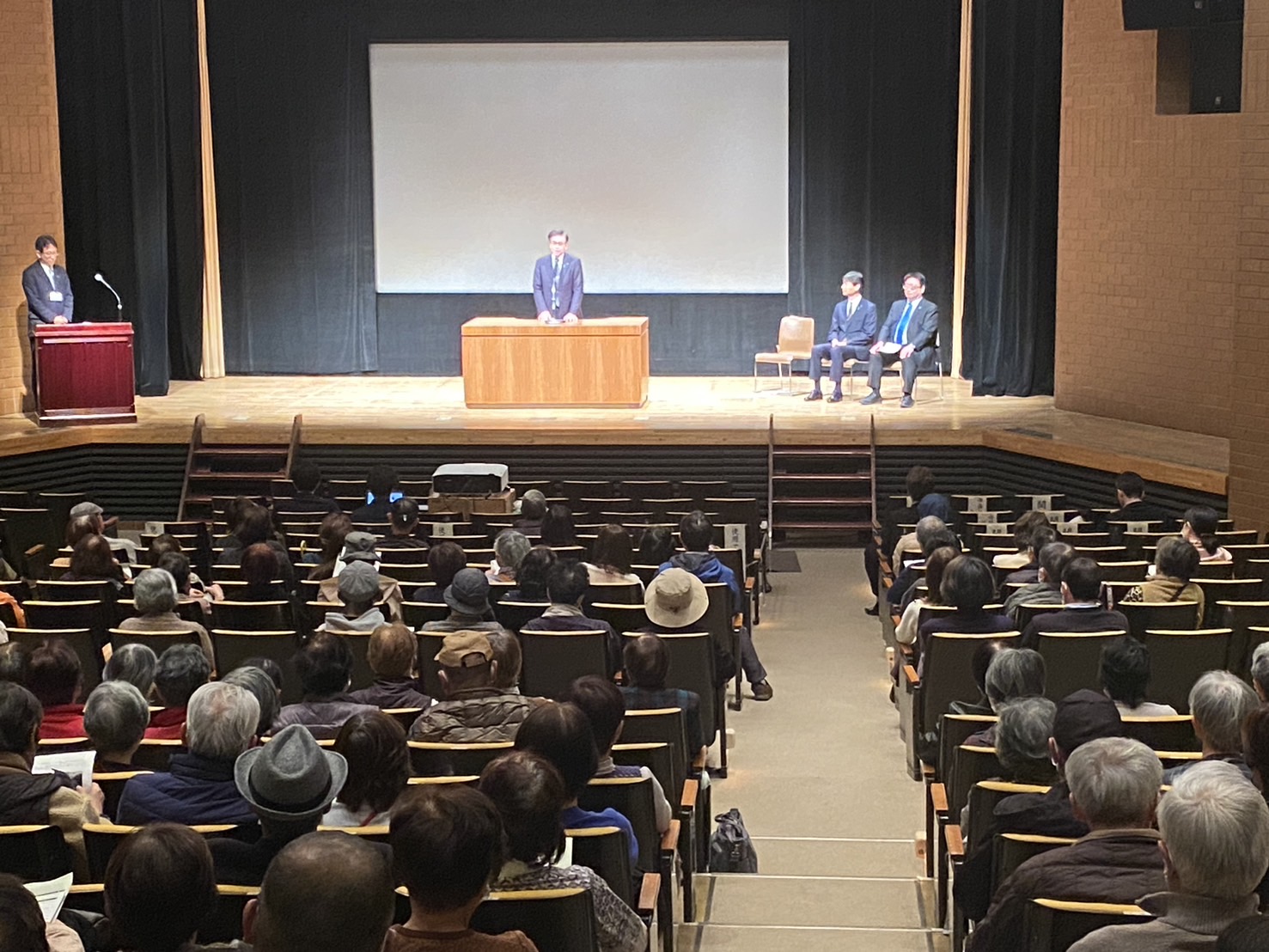 満席の観客が見守る中、舞台上の演壇で市長が挨拶し、関係者が横に座るホールでの式典全体風景の写真