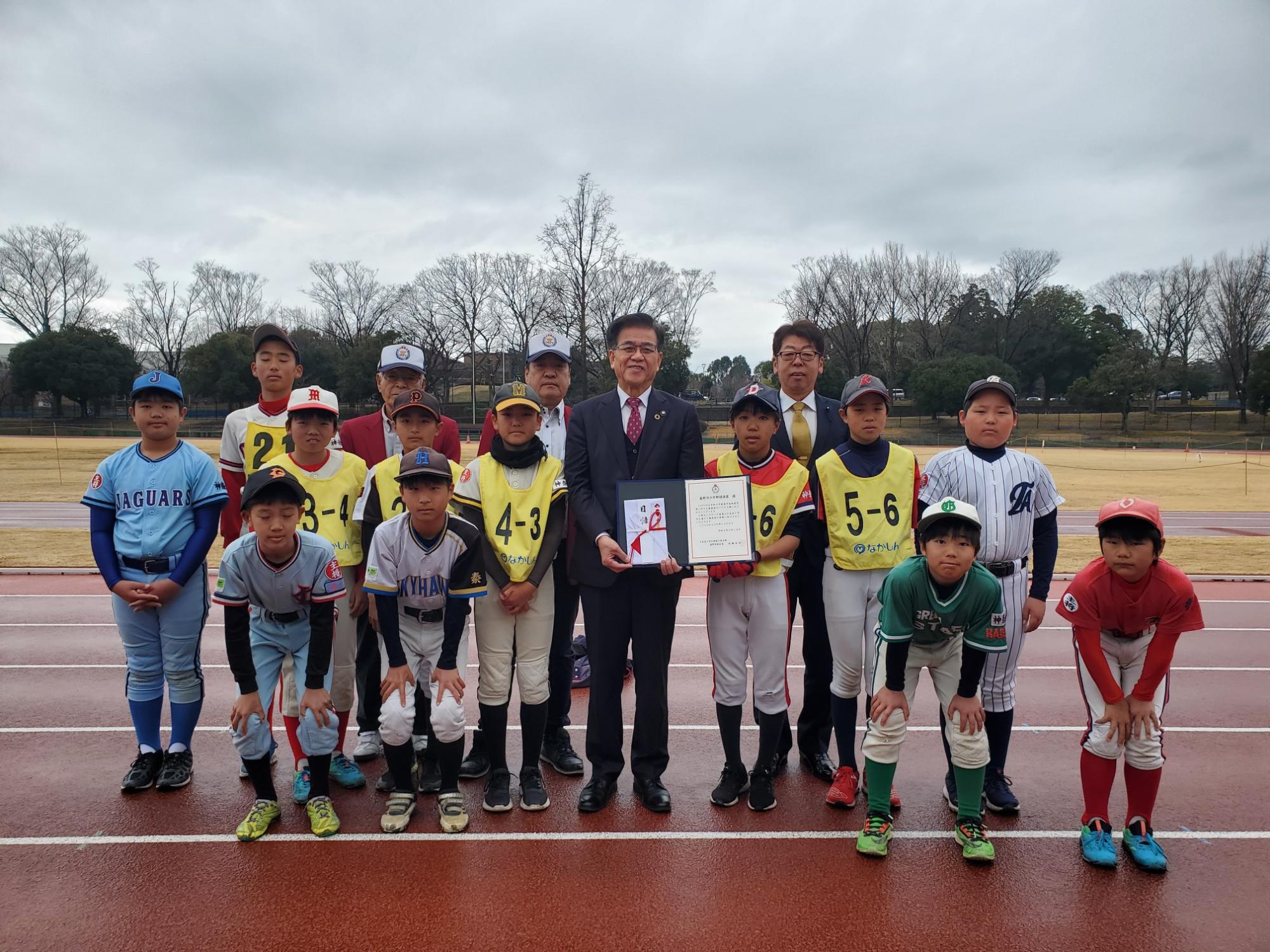 複数のチームユニフォームを着た少年野球選手たちが整列し、中央の市長が賞状を持って記念撮影している屋外集合写真