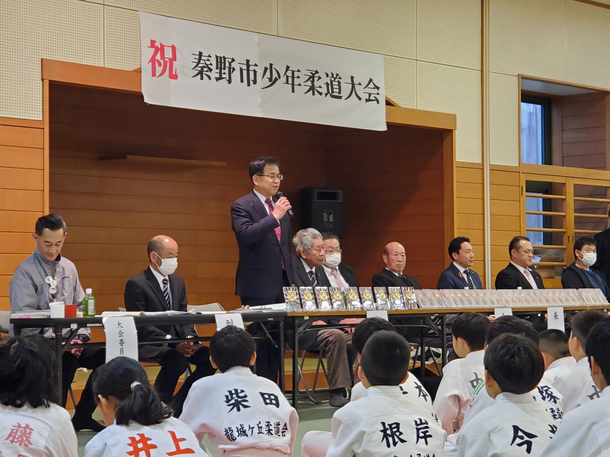 柔道着を着た子どもたちが正座して壇上の挨拶を聞き、背後の長机には大会関係者と多数の賞牌が並ぶ柔道大会会場の写真