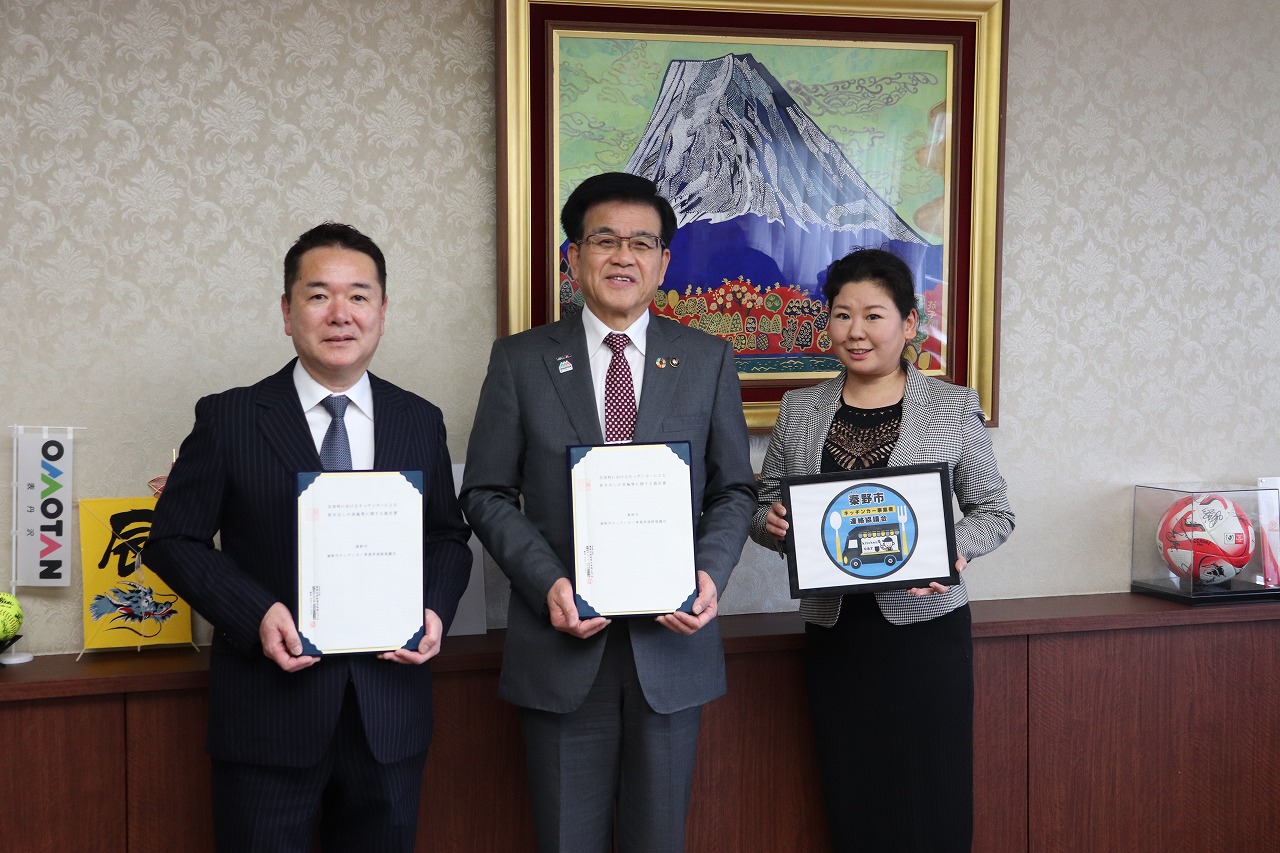 中央の市長と左の男性は協定書のようなものを手に持ち、右の女性は「愛野市 公共交通応援団」と書かれたバスのデザイン入りのパネルを掲げて記念撮影している写真