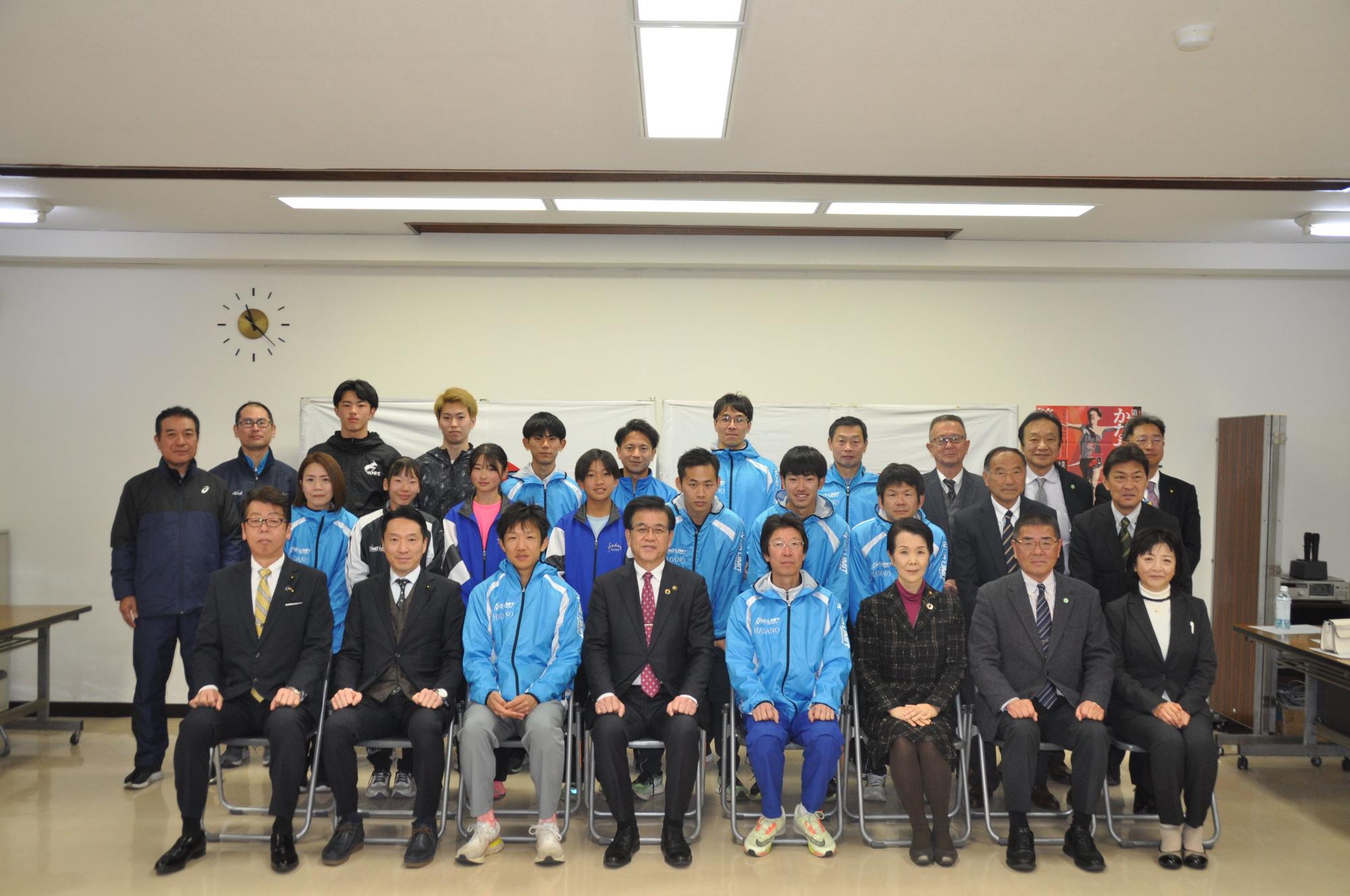 会議室で青いジャンパー姿の若者たちとスーツ姿の関係者が数列に並び、市長を中心に集合写真を撮影している写真