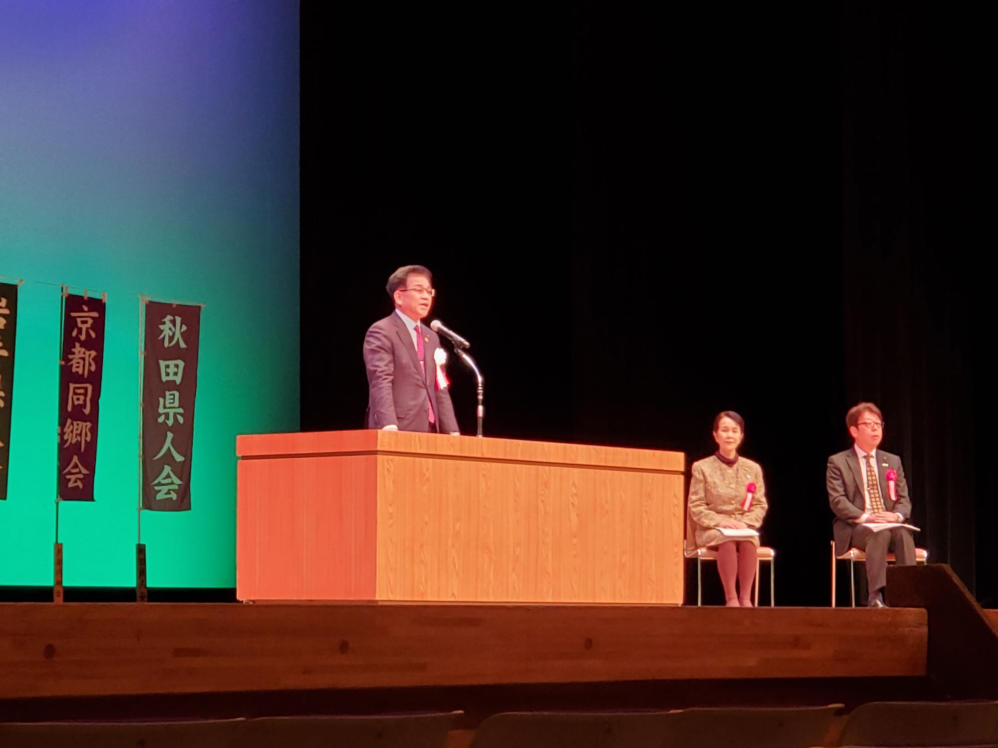 演台でスーツ姿の市長が演説し、背後に「秋田県人会」などの旗と椅子に座る2人の来賓の方々の舞台上の写真