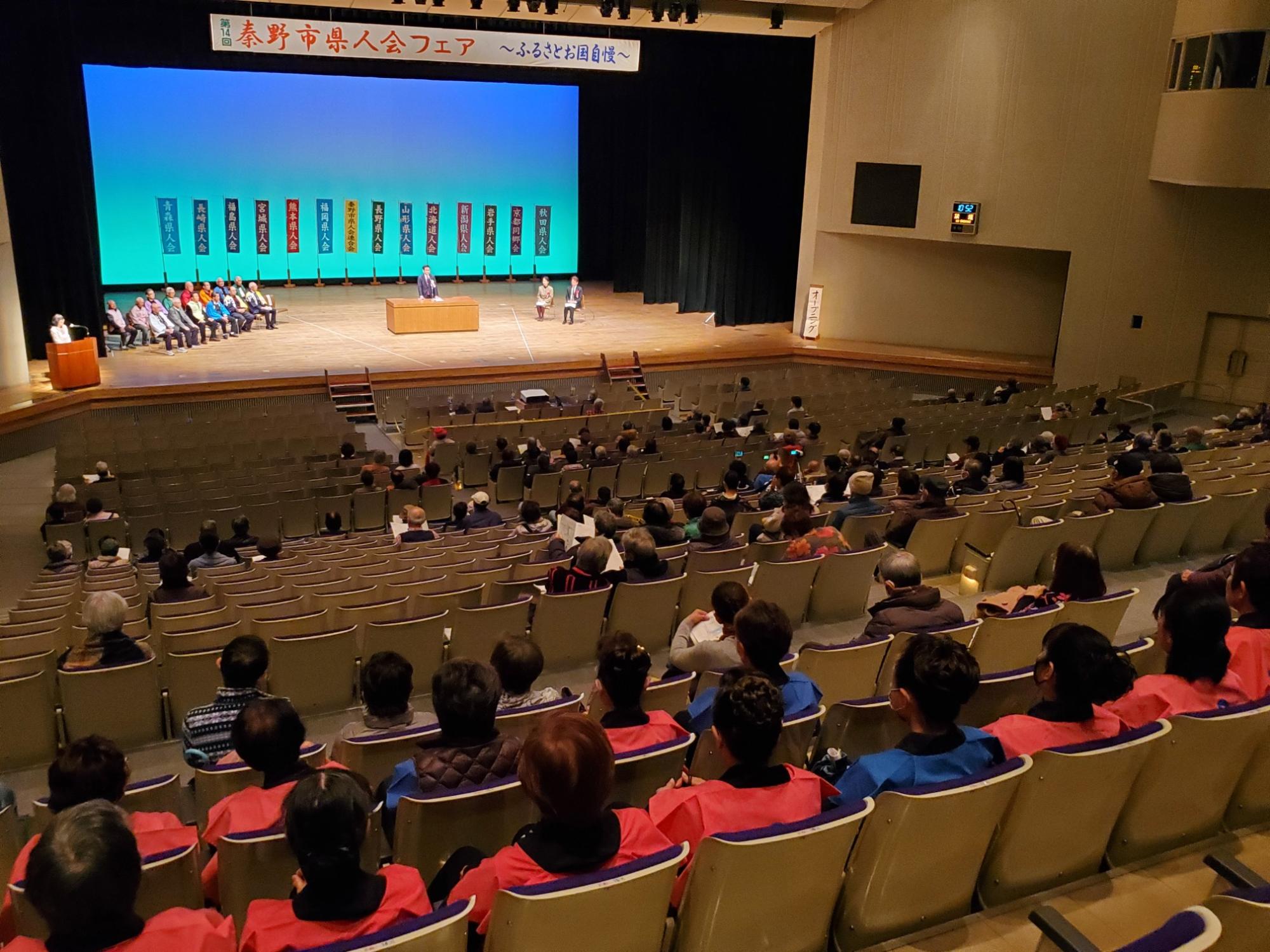 大きなホールで「秦野市県人会フェア」と書かれた横断幕の下、壇上で演説が行われ観客が多数着席している様子を後方から写した写真