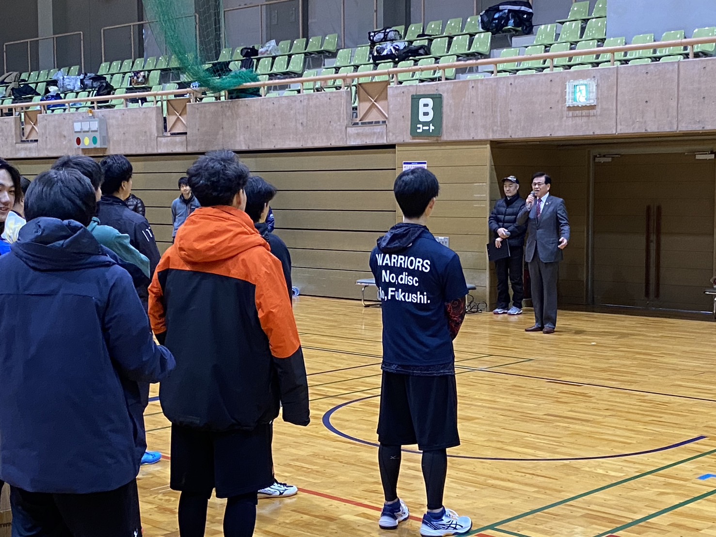 体育館のコートでスーツ姿の市長がマイクを持ち挨拶し、競技用ウェア姿の選手の方々が集まって聞いている様子の写真