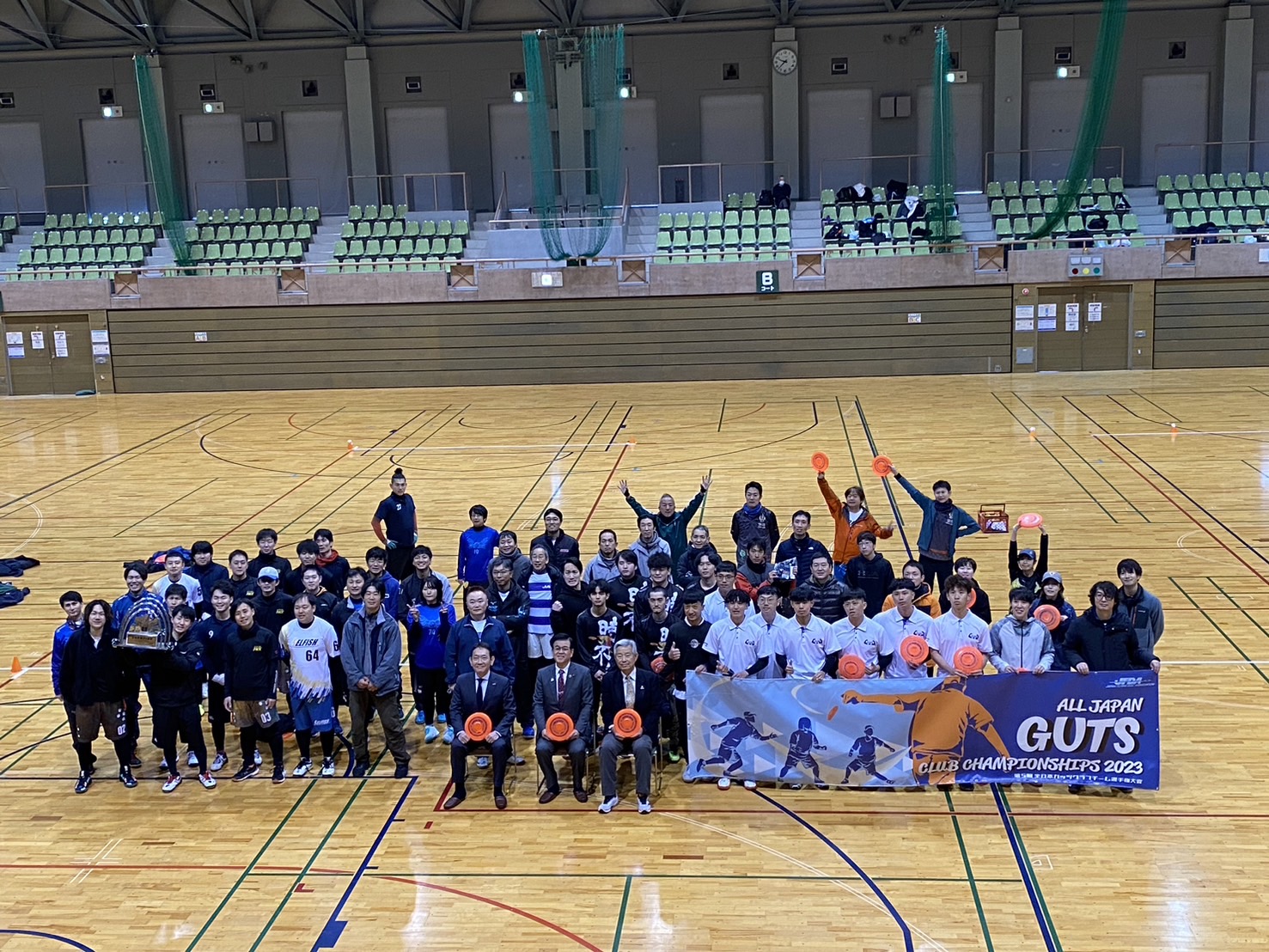 体育館のコート中央で多数の参加者が並びフライングディスクを掲げ、横断幕を前に記念撮影している大会集合写真