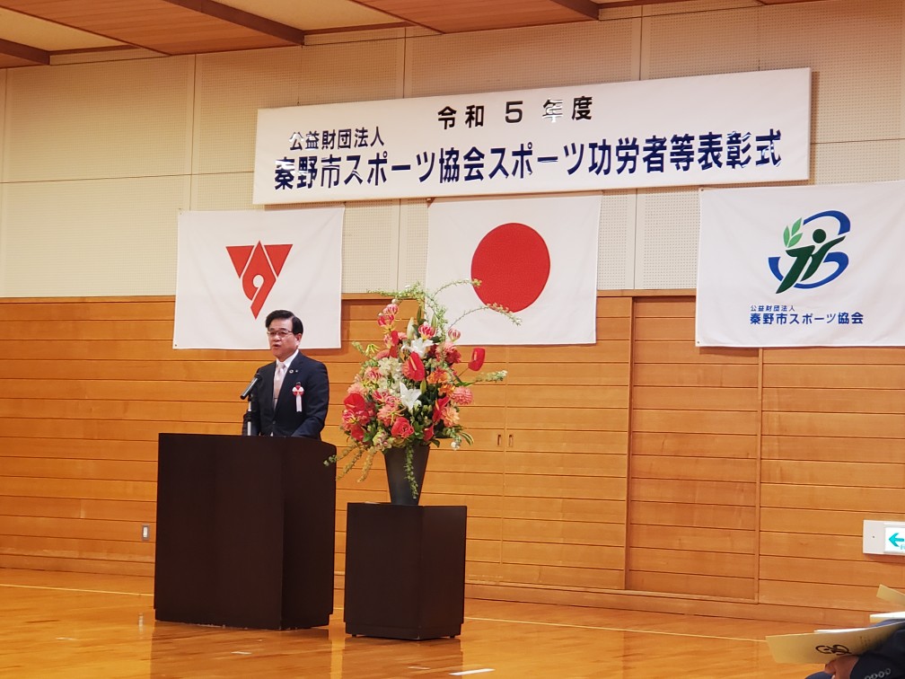壇上でスーツ姿の市長が演説しており、背後には日本国旗と横断幕が掲げられ花が飾られた表彰式会場の写真