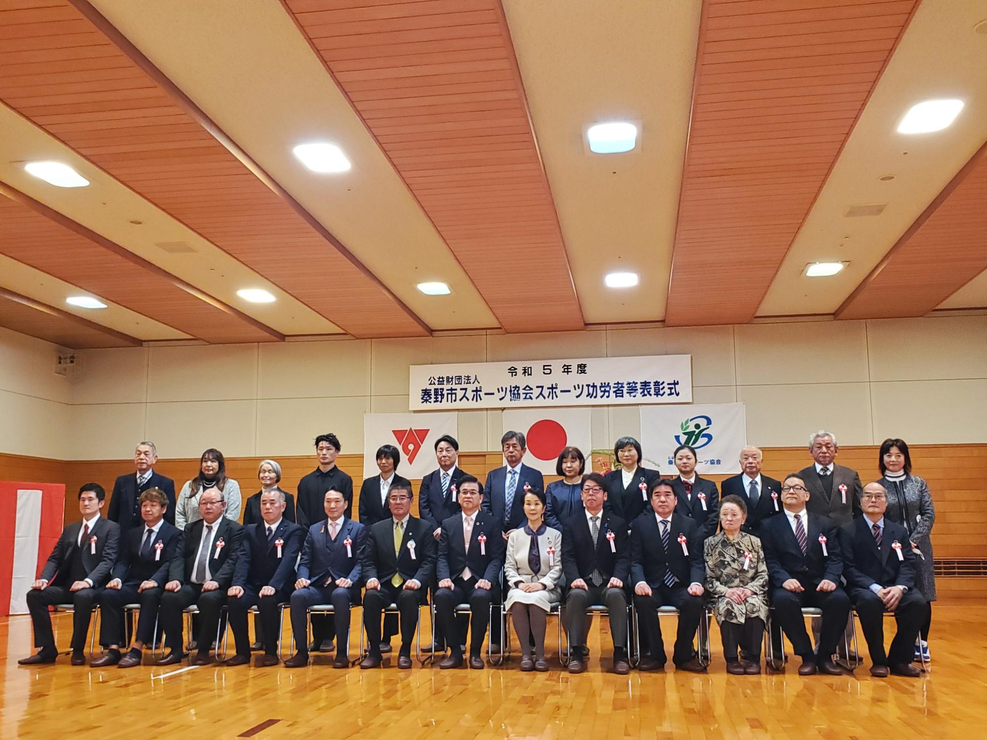 表彰式の会場で受賞者や関係者が2列に整列して椅子に座り、背後に横断幕と日本国旗が掲げられた集合写真