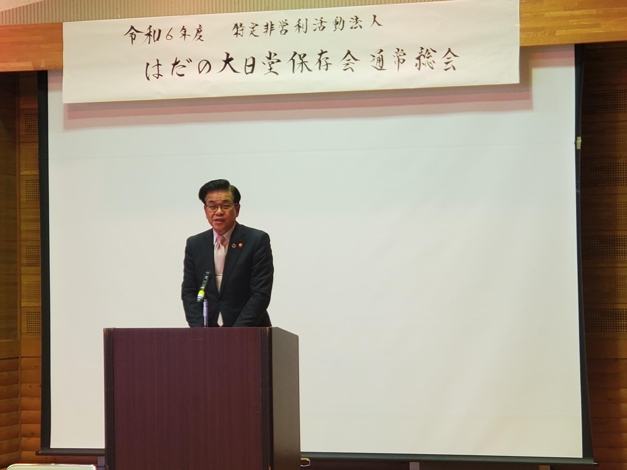 スーツ姿の市長が演台に立ちマイクで発言しており、背後には令和6年度はだの大日堂保存会通常総会と書かれた横断幕が掲げられた会場の写真
