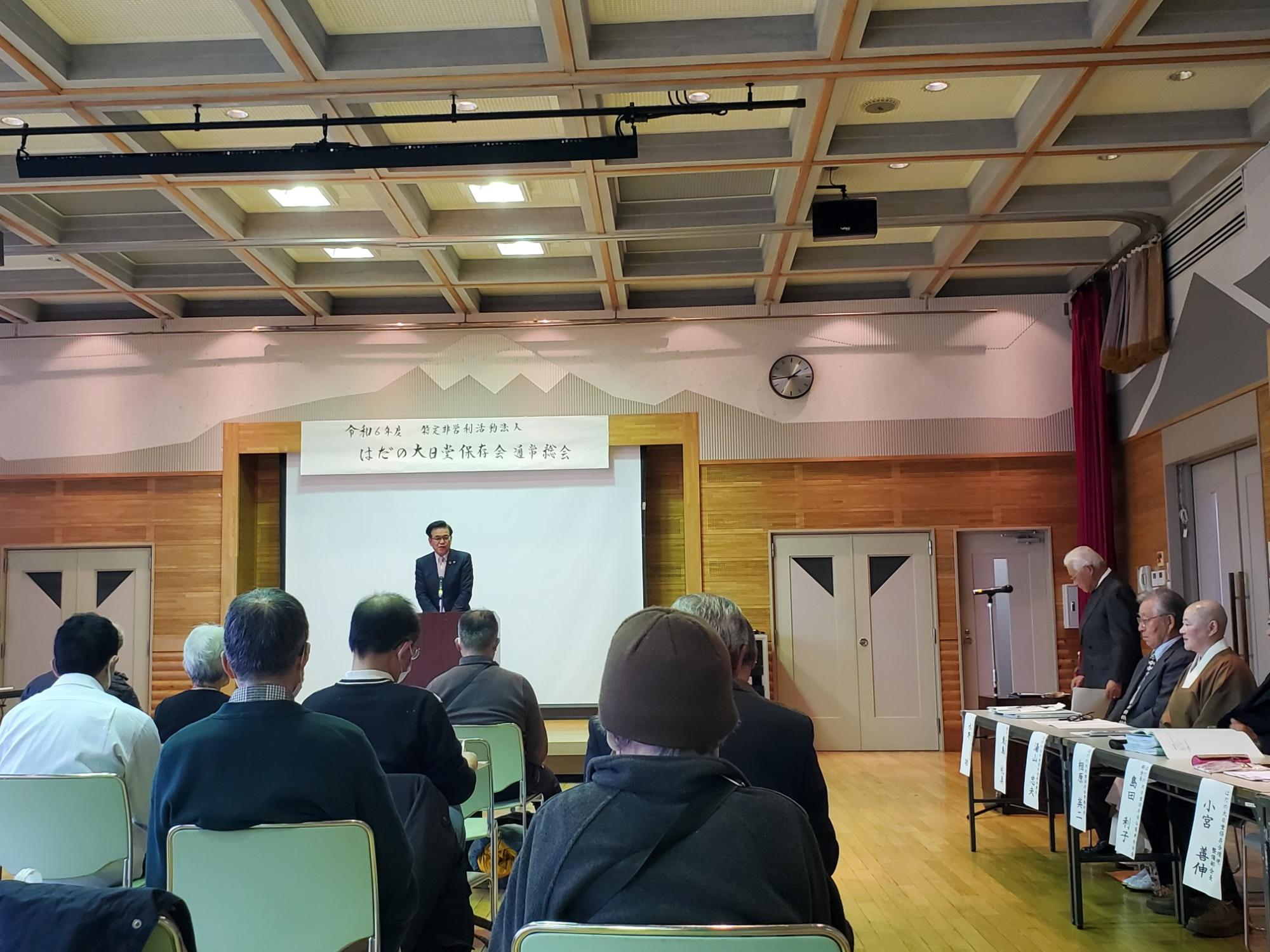 壇上で市長が演説し正面には多くの聴衆が座り、右側には役員らが並ぶ通常総会の会場の写真