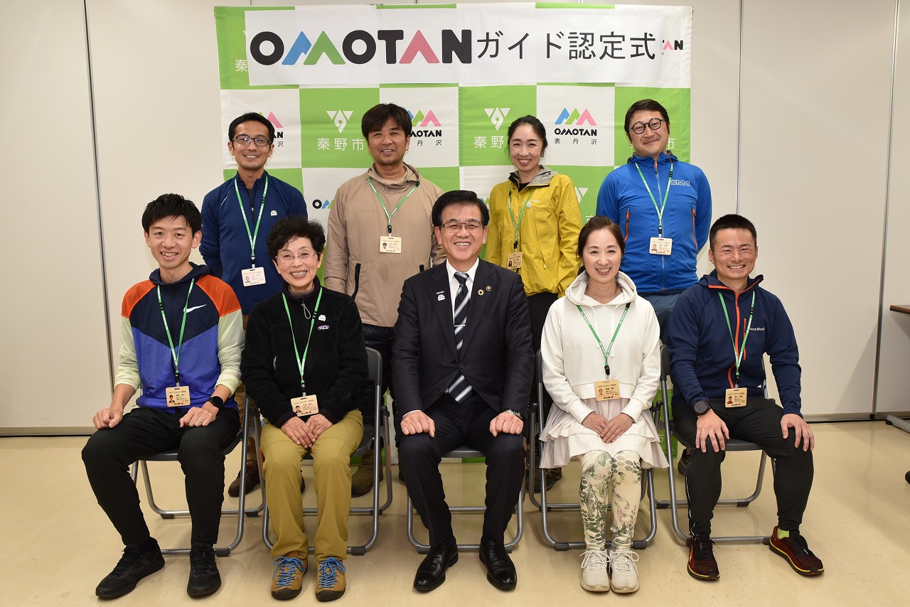 緑色の認定証パネルを背景に、スーツ姿の市長と首から認定証を下げた男女が二列に並び笑顔で撮影している集合写真