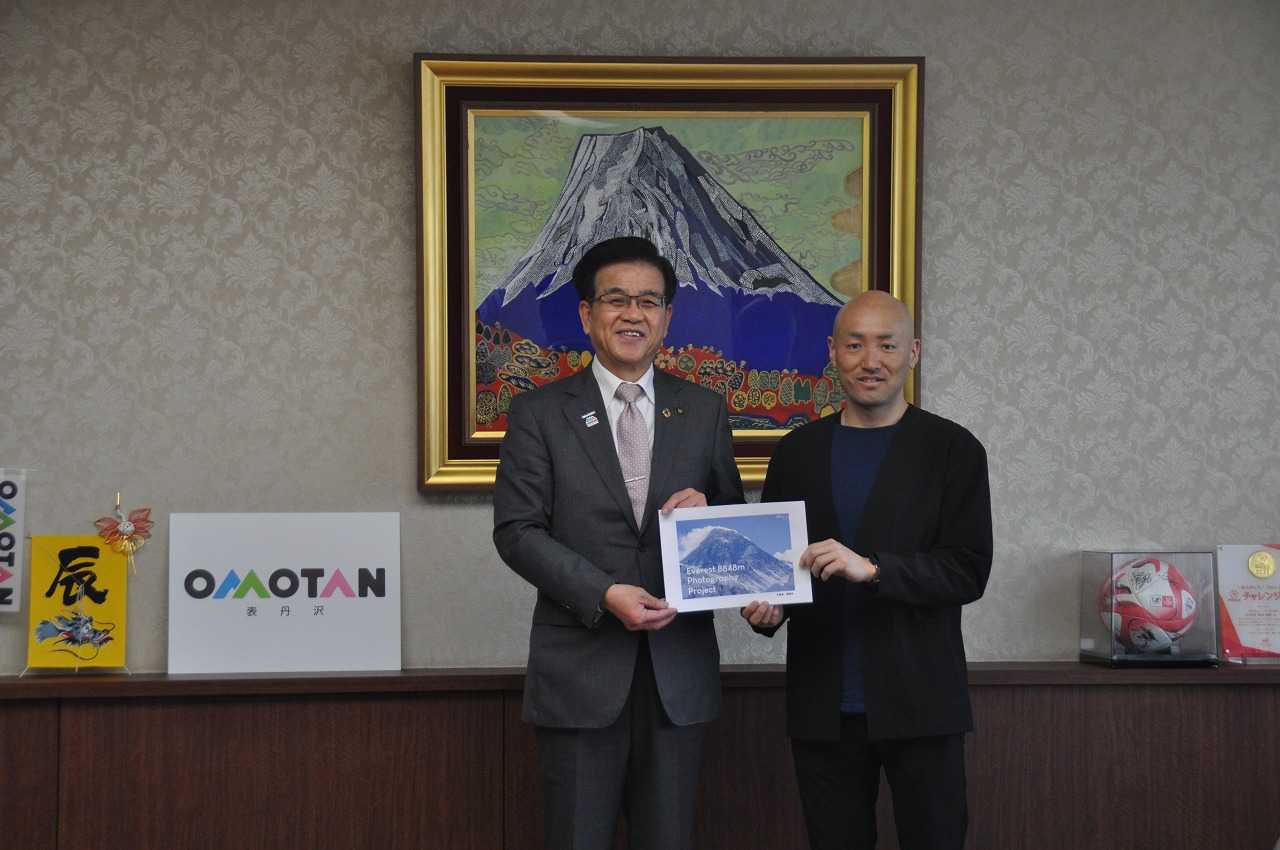 スーツ姿の市長と黒い服装の関健作が並び、富士山写真のパネルを一緒に持っている記念写真