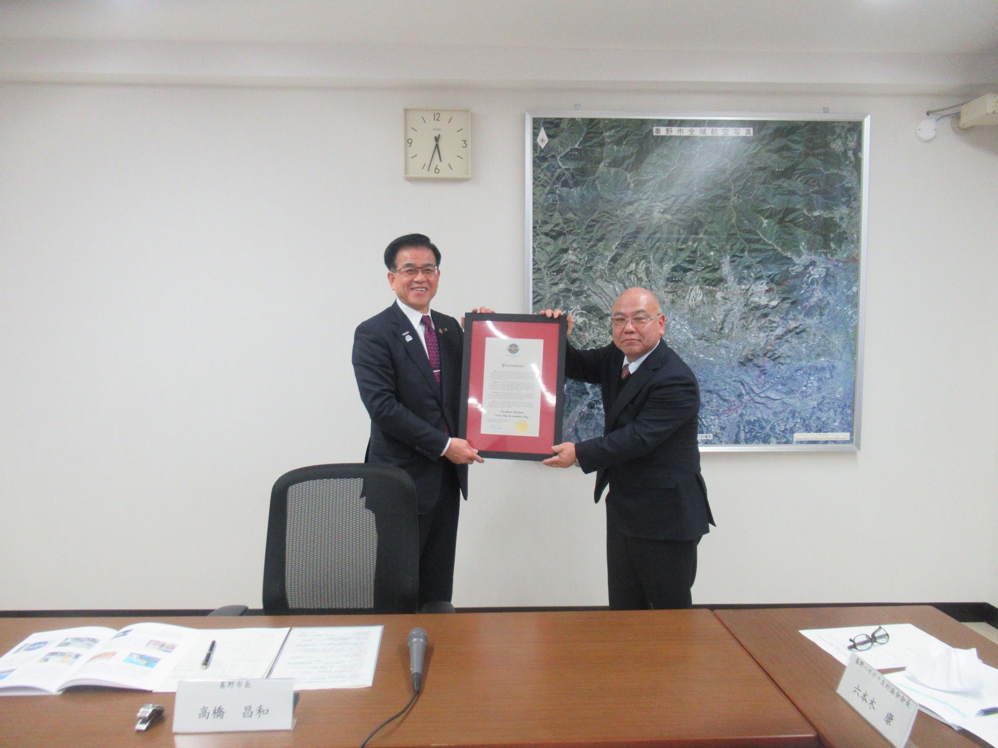 市長と青少年訪問団の代表の方が会議室で額縁に入った表彰状を笑顔で持ち記念撮影している写真