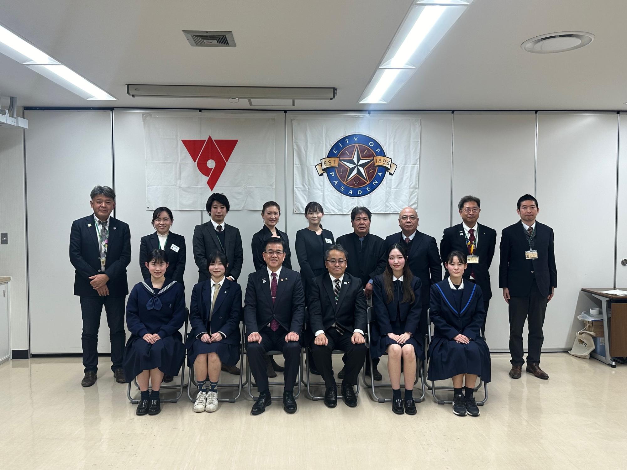室内でスーツ姿の市長や関係者と制服姿の学生が二列に並び、背後に市章やロゴの旗を掲げて記念撮影している写真