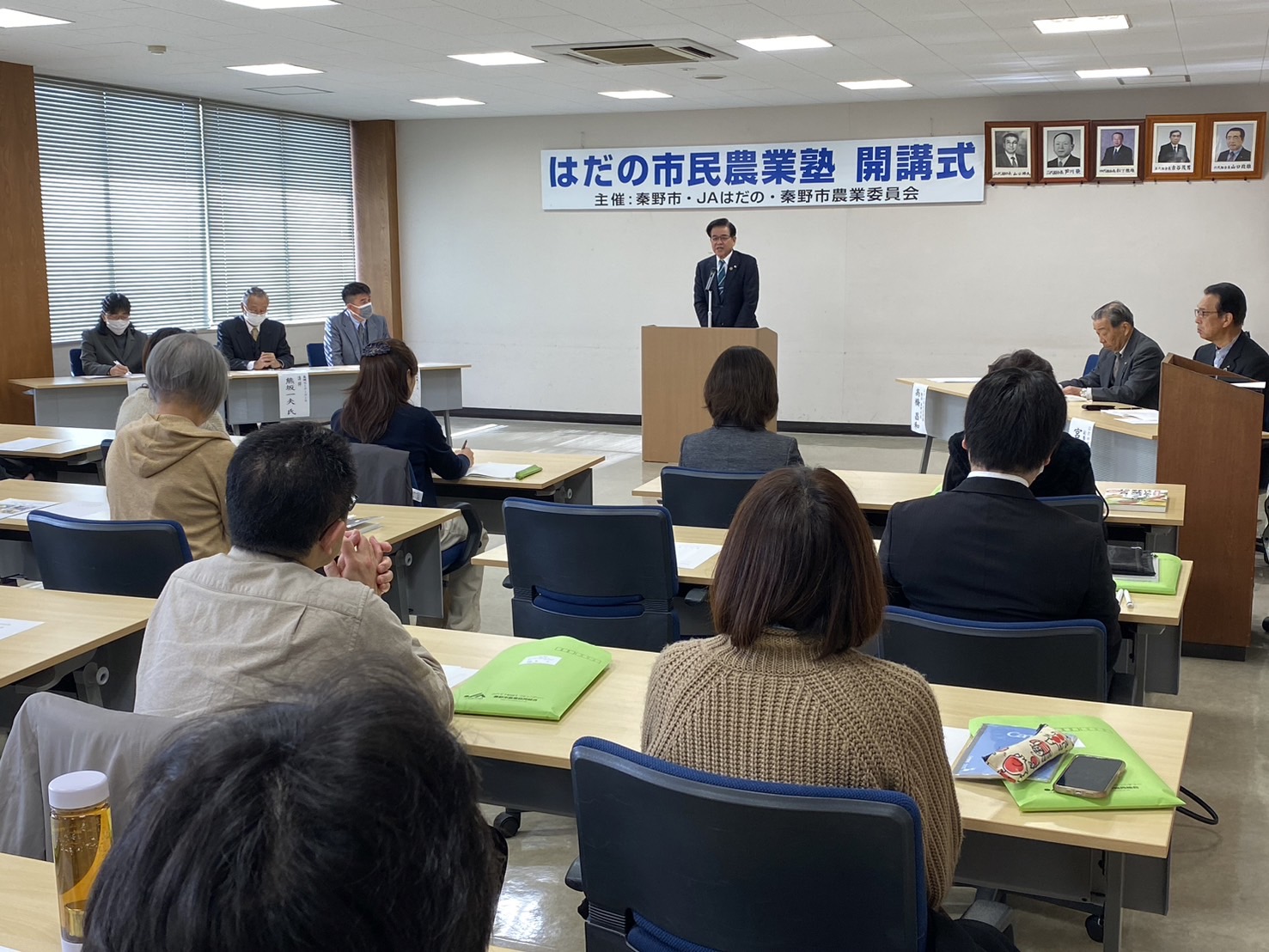 「市民農業塾 開講式」の横断幕がある室内で、スーツの市長が挨拶し、机に向かって座る大勢の参加者が聞いている様子を後方から写した写真