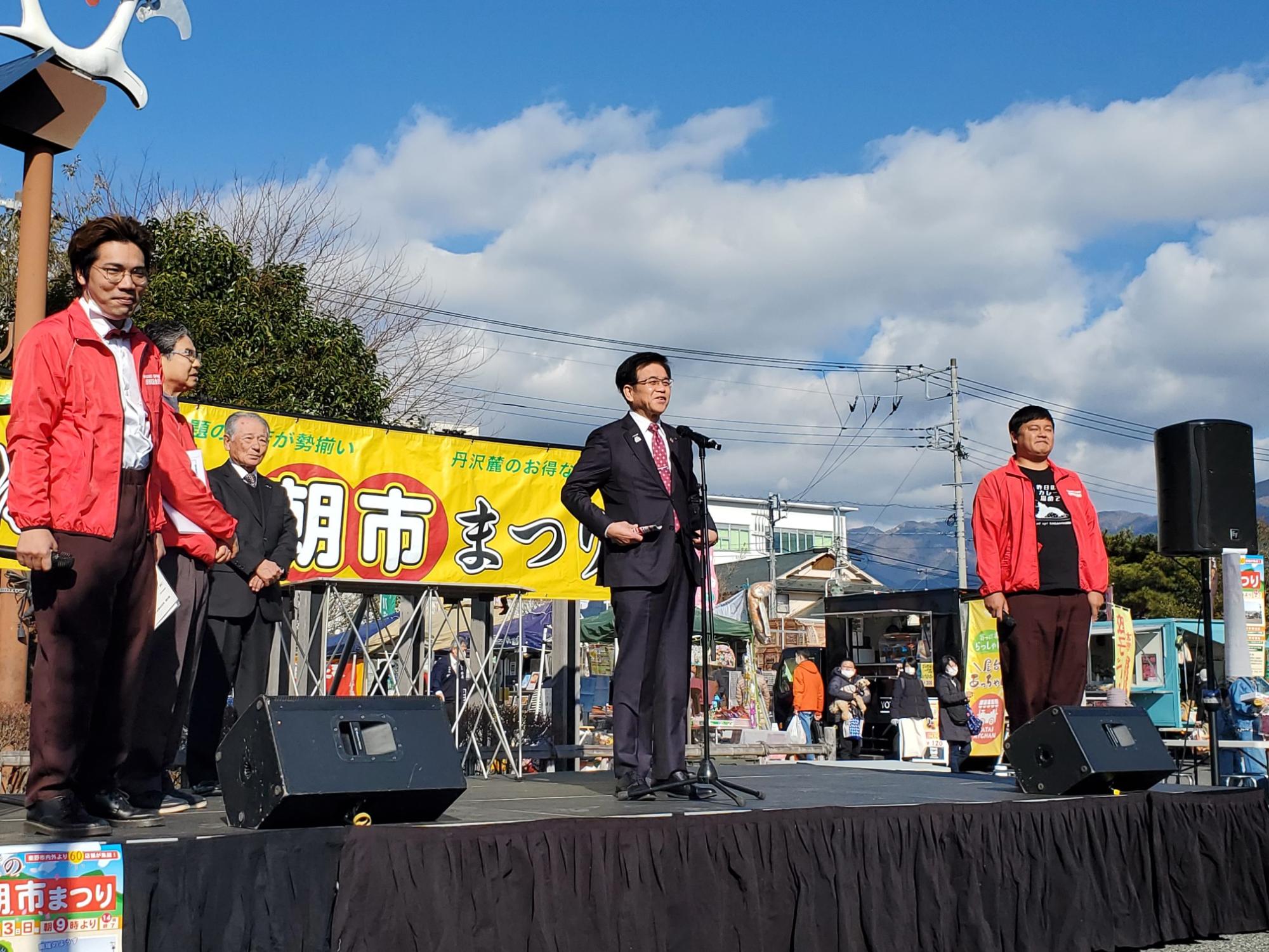 「市まつり」と書かれた黄色い横断幕を背景に、スーツ姿の市長が屋外ステージのマイクの前で挨拶している写真