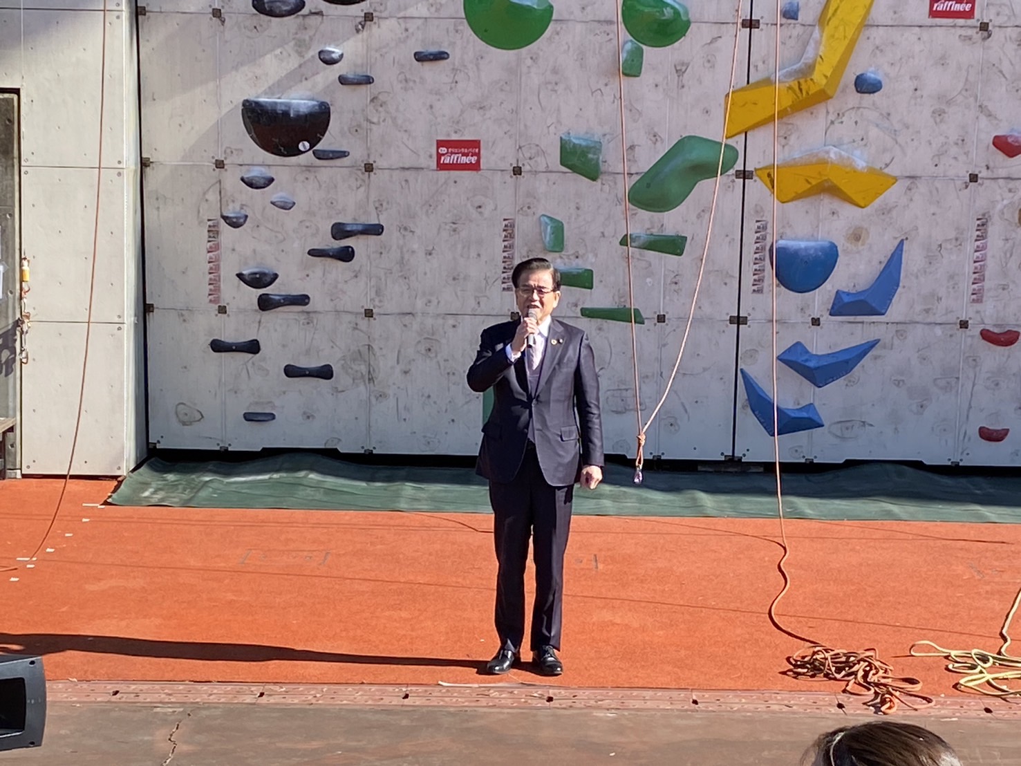 そびえ立つクライミングウォールを背景に、スーツ姿の市長がマイクを手に持ち、イベントの開会式で挨拶している写真