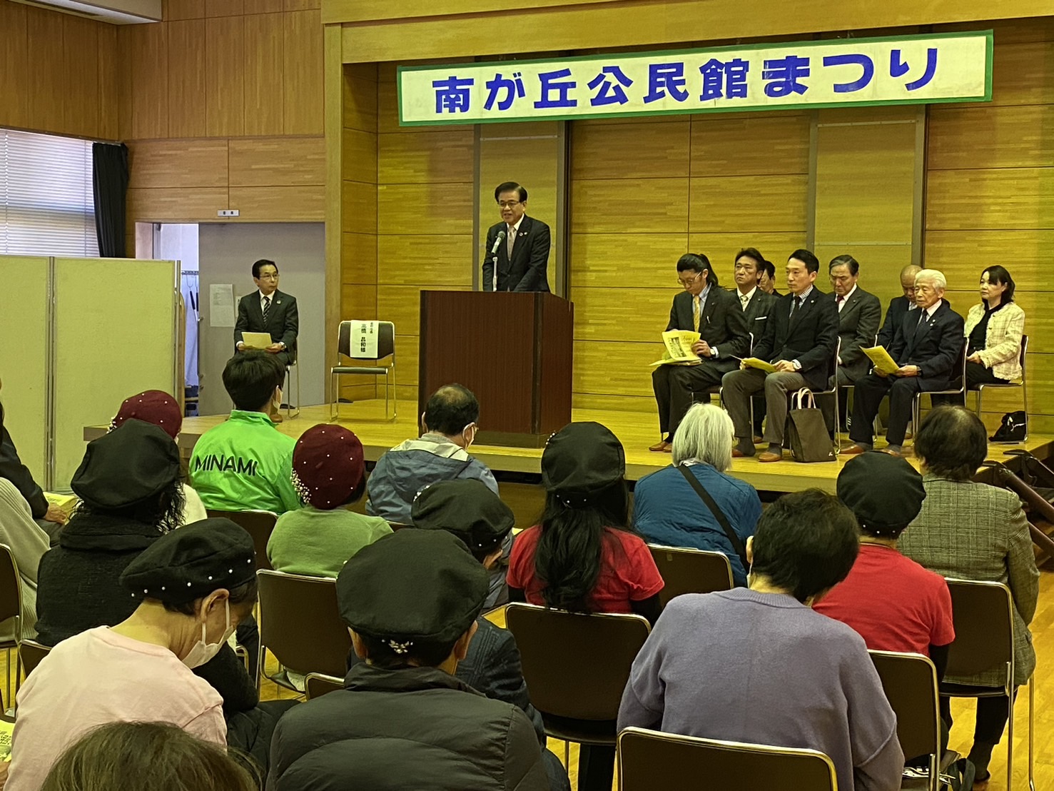 「南が丘公民館まつり」と書かれた横断幕の下、ステージでスーツの市長が挨拶し、壇上の来賓と客席の観客が聞いている様子の写真