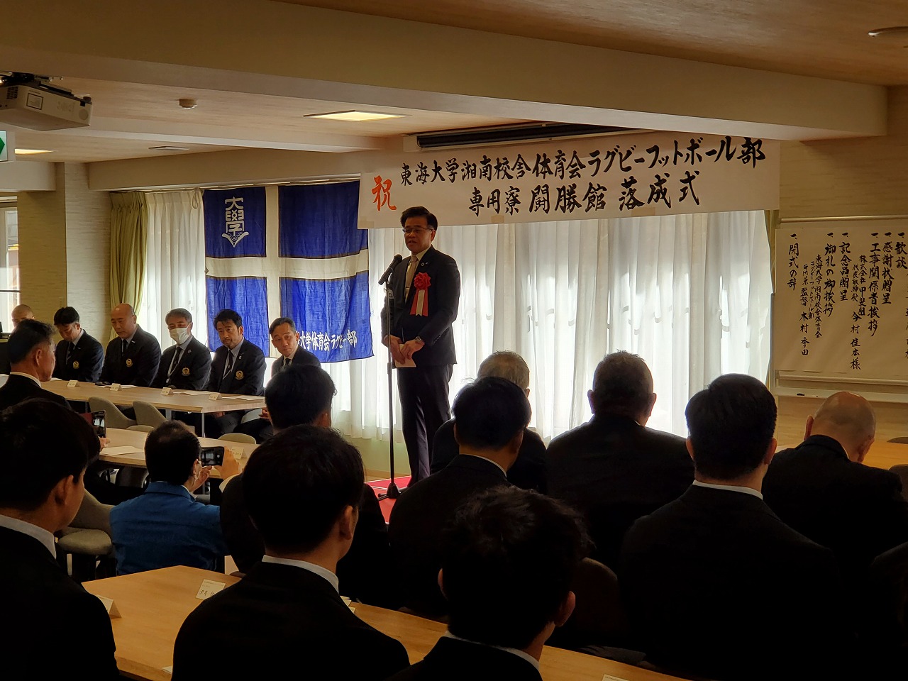 ラグビー部の「専用開寮・開藤館 落成式」と書かれた横断幕の前で、スーツの市長が挨拶し、多くの出席者が聞いている写真