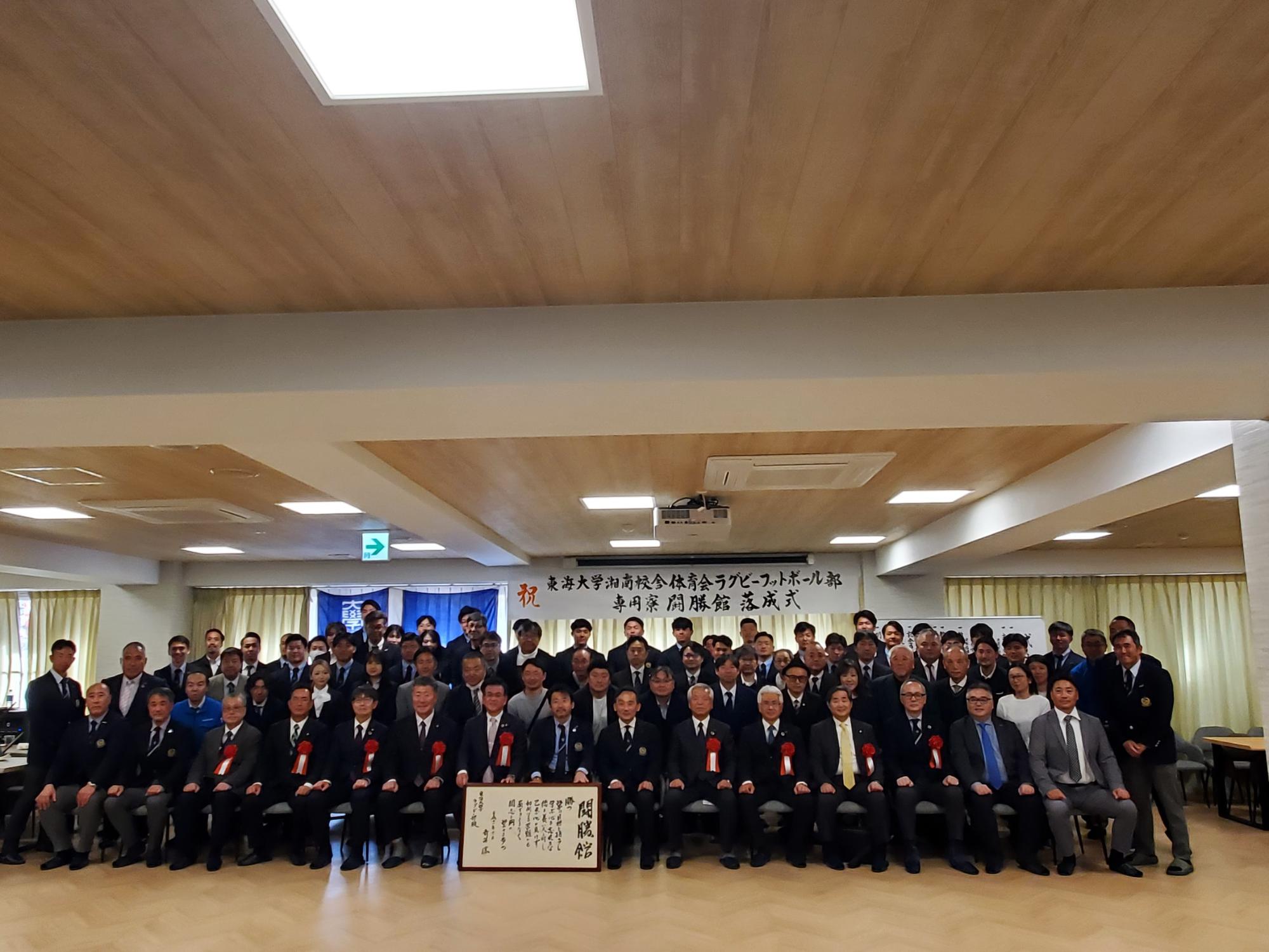 「ラグビーフットボール部 落成式」と書かれた横断幕を背景に、スーツ姿の大勢の参加者が集合写真を撮っている写真