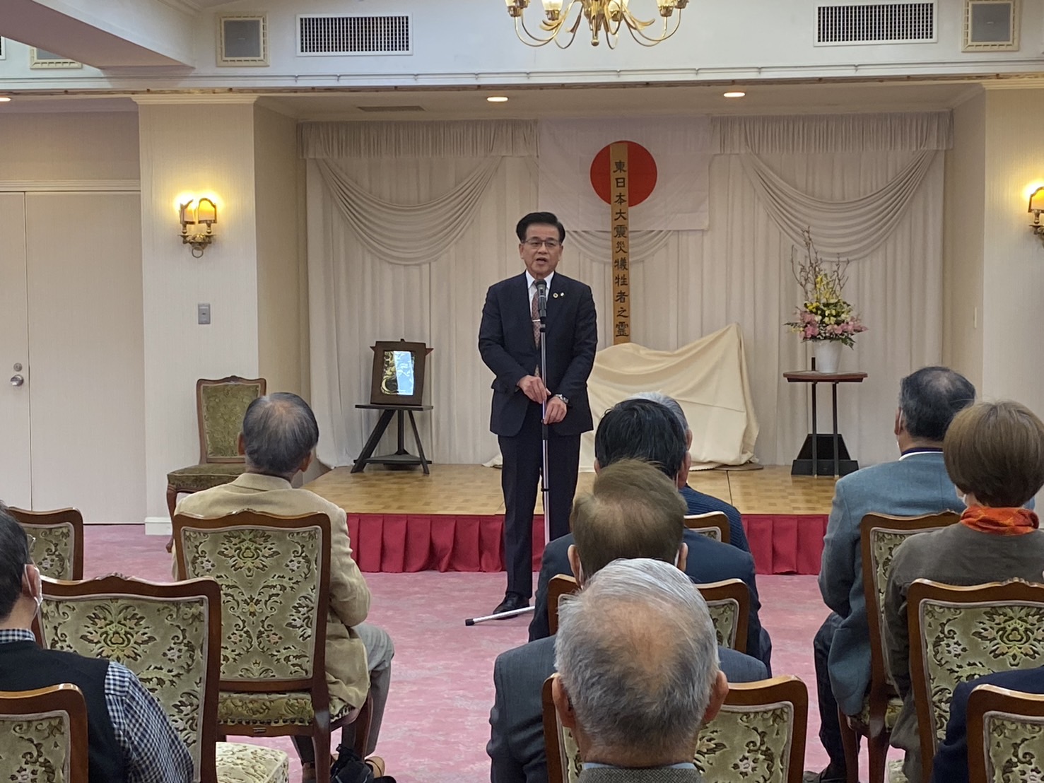 「東日本大震災犠牲者之霊」と書かれた幕の前で、スーツの市長がマイクに向かって挨拶し、椅子に座った参列者が聞いている様子の写真