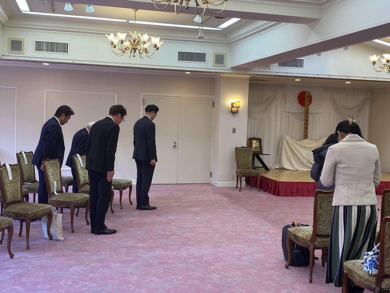 スーツ姿の参列者たちが一斉に起立し、深く頭を下げて黙祷を捧げている追悼式の写真