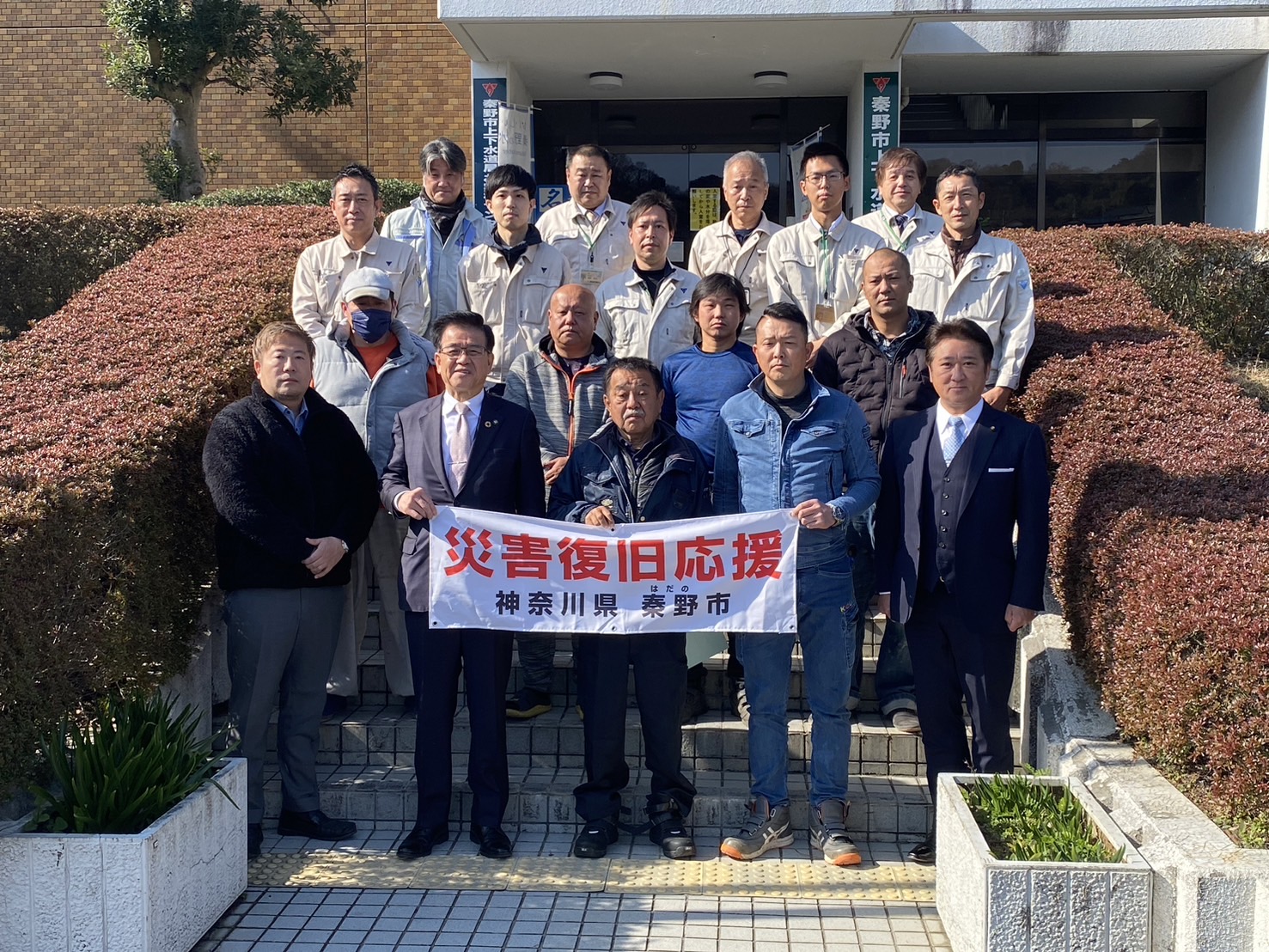 「災害復旧応援」の横断幕を、スーツや作業着姿の男性たちが建物の前の階段で持ち、記念撮影している写真