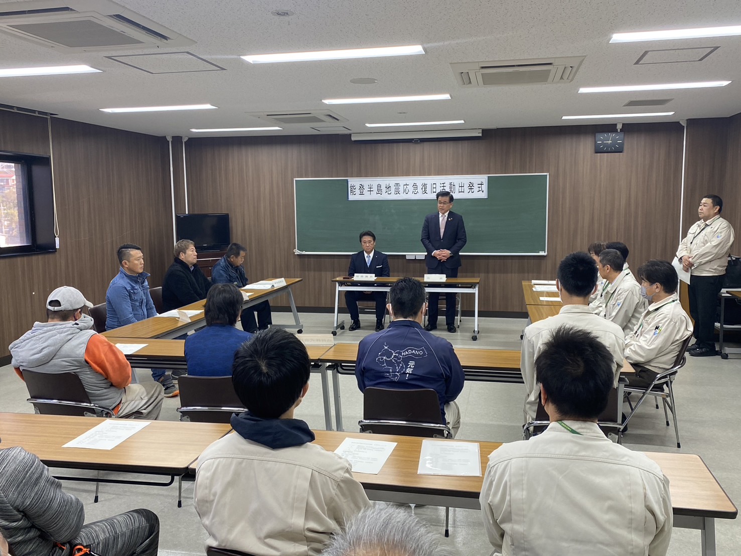 「出発式」と書かれた黒板を背に、机に向かって座る作業着姿の隊員たちに向かってスーツの市長が話をしている写真