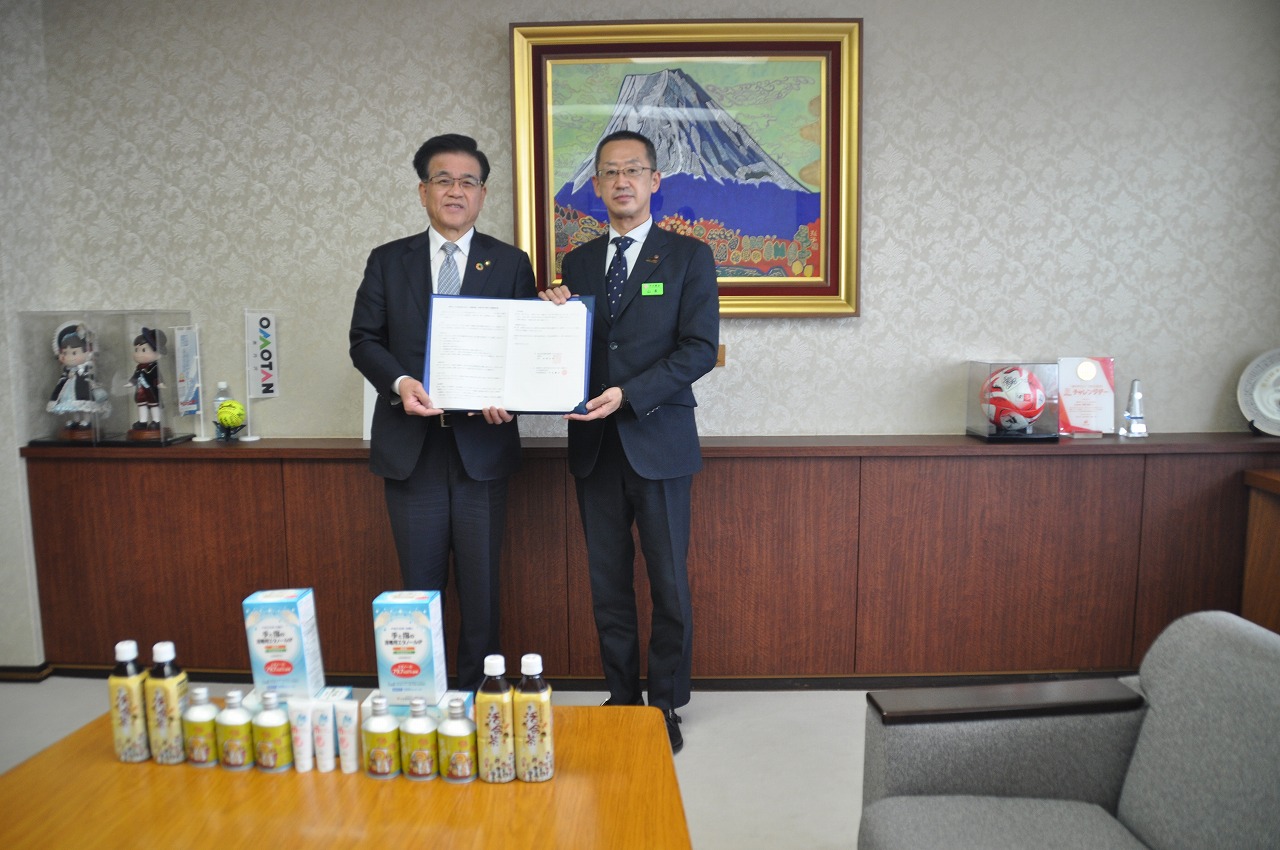 スーツ姿の市長と中北薬品の方が協定書のようなものを一緒に手に持ち、テーブルに並べられた商品を前に記念撮影をしている写真