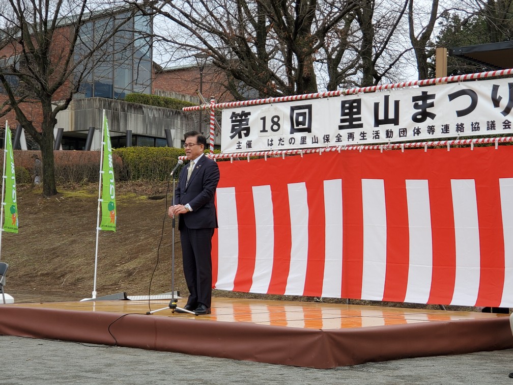 「第18回 里山まつり」と書かれた横断幕が掲げられた屋外ステージで、紅白幕を背景にスーツ姿の市長がマイクスタンドの前に立ち挨拶している写真