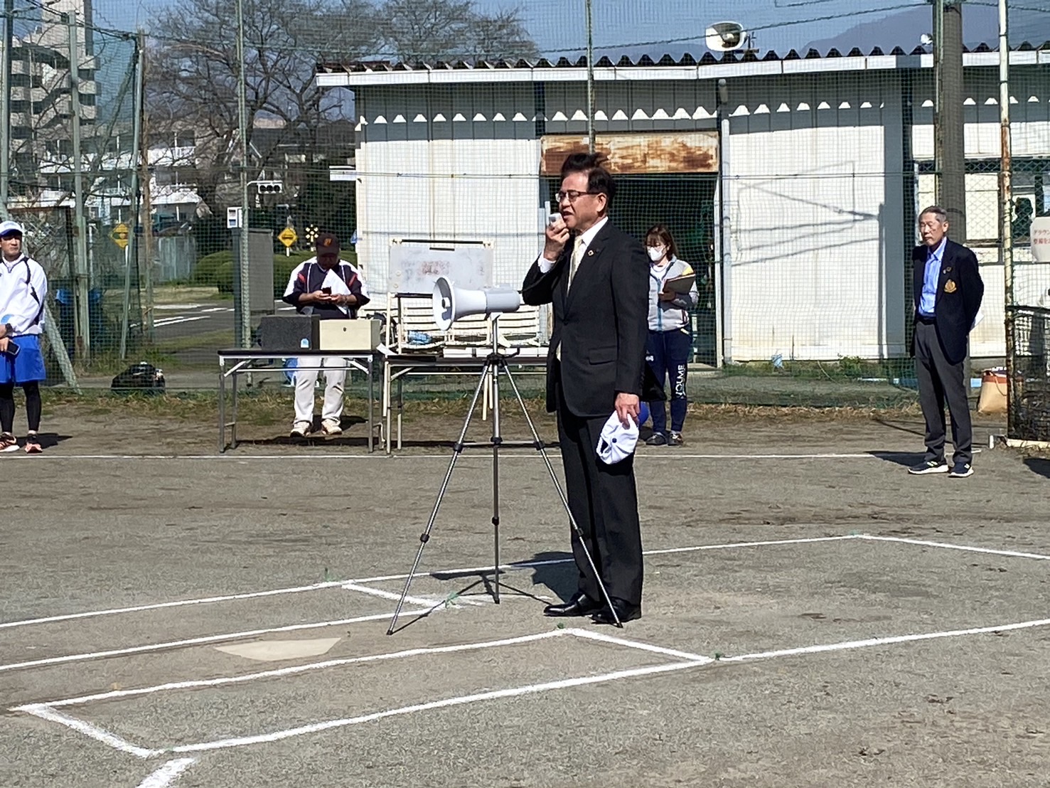 グラウンドのホームベース付近にスーツ姿の市長が、三脚に設置されたメガホンに向かって開会式の挨拶をしている写真