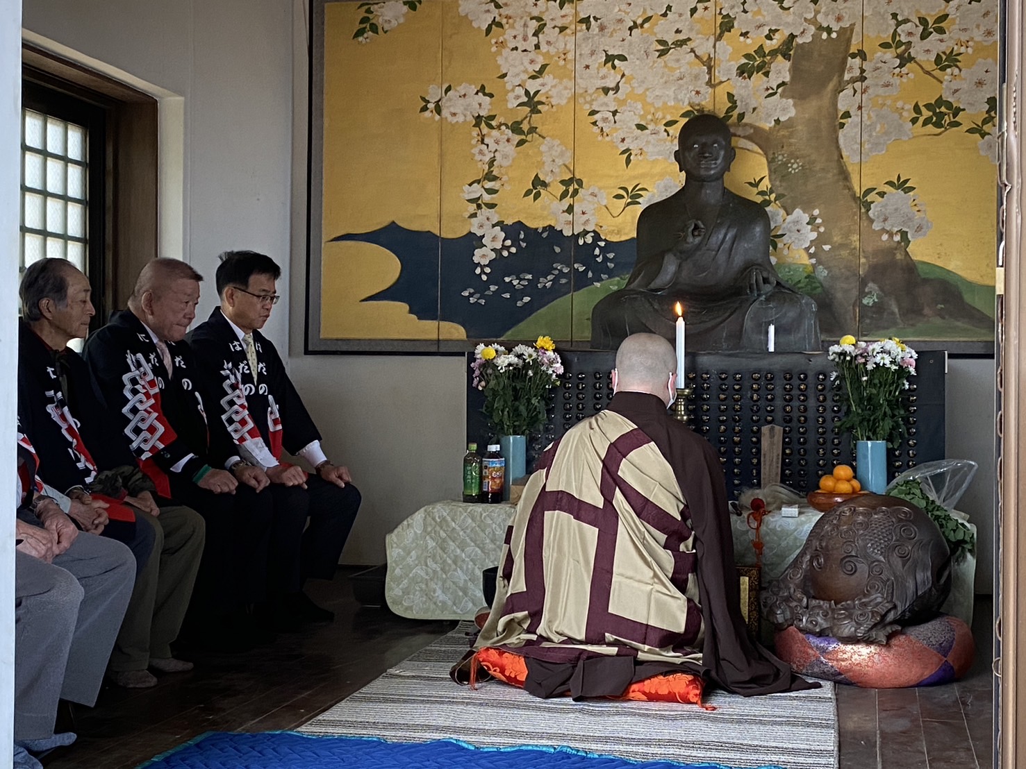 金屏風の前の仏像に向かい、袈裟姿の僧侶が供養を行い、それを法被を着た関係者の方々が横に座って静かに見守っている写真