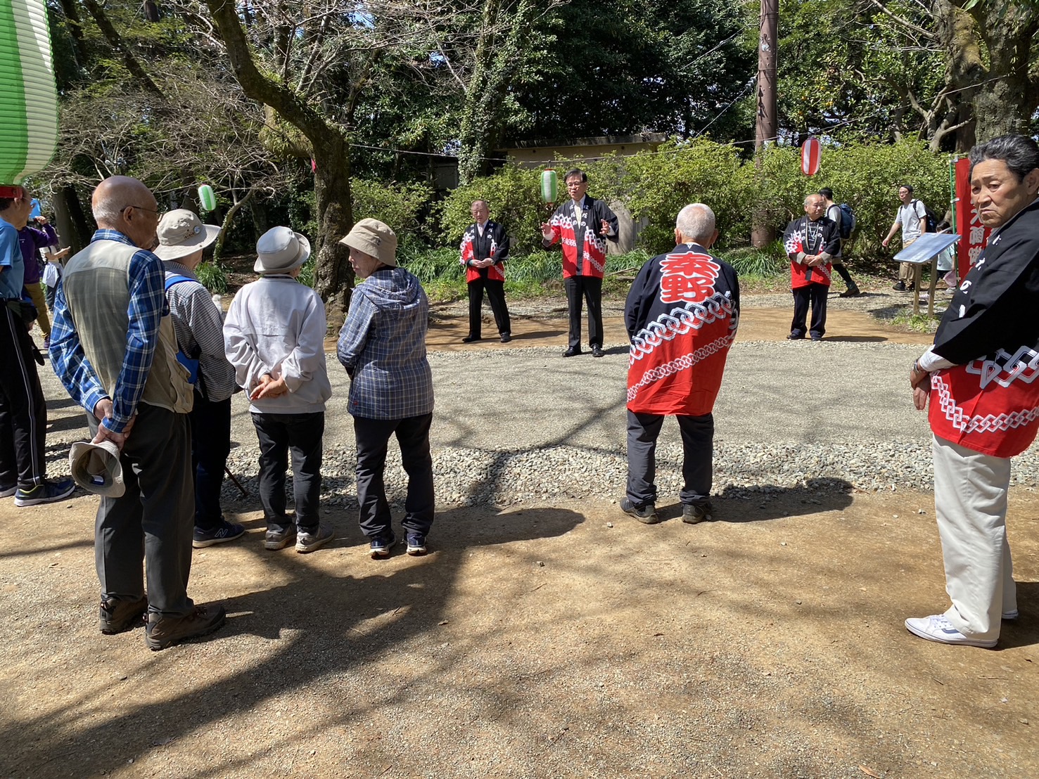 緑豊かな屋外で、法被を着た市長が挨拶している様子を、ハイキングの服装をした数名の方々が輪になって聞いている写真