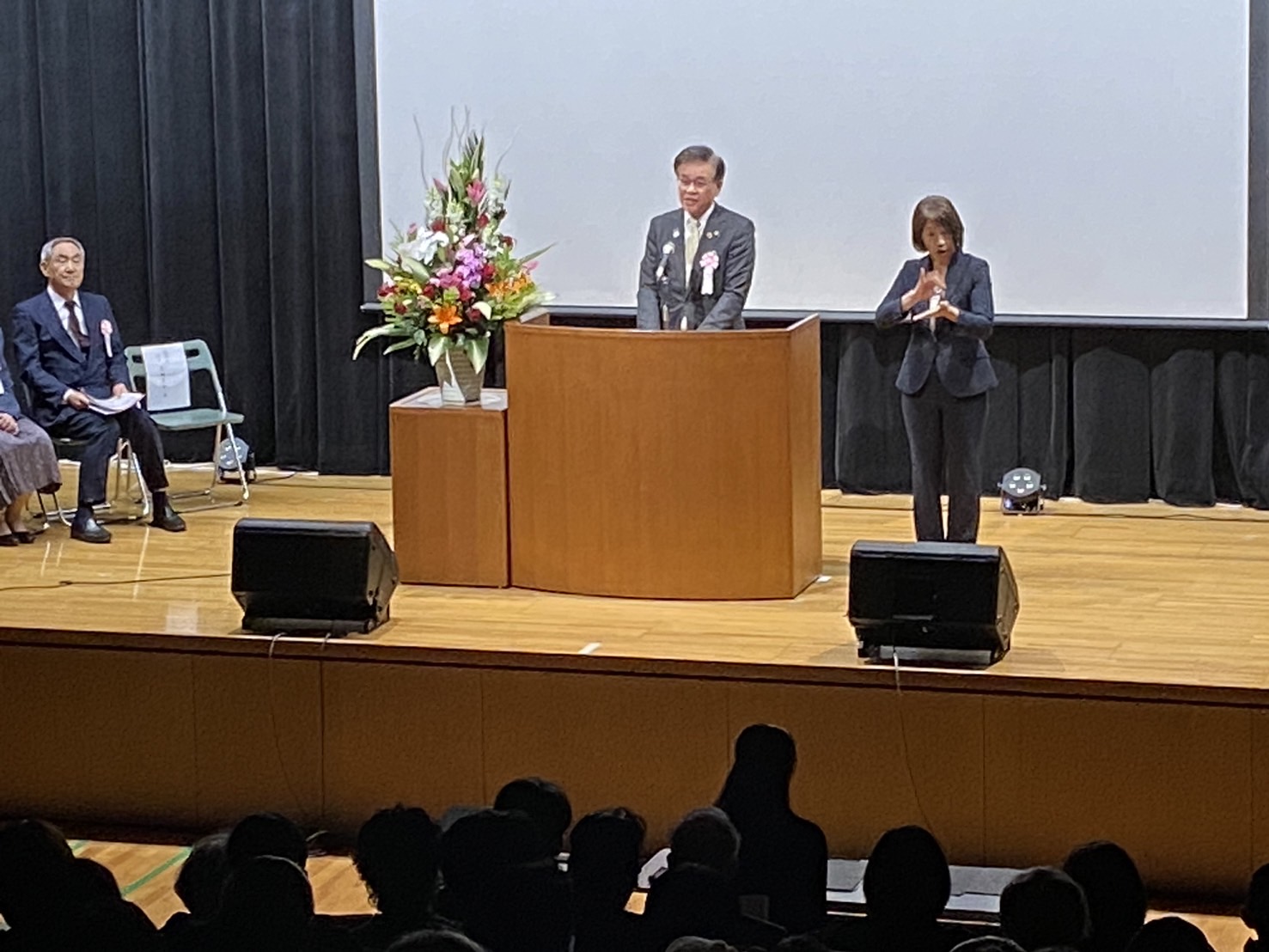 ステージ上の演台で、スーツ姿の市長がスピーチし横に立つ女性が話に合わせて手話をする様子の写真