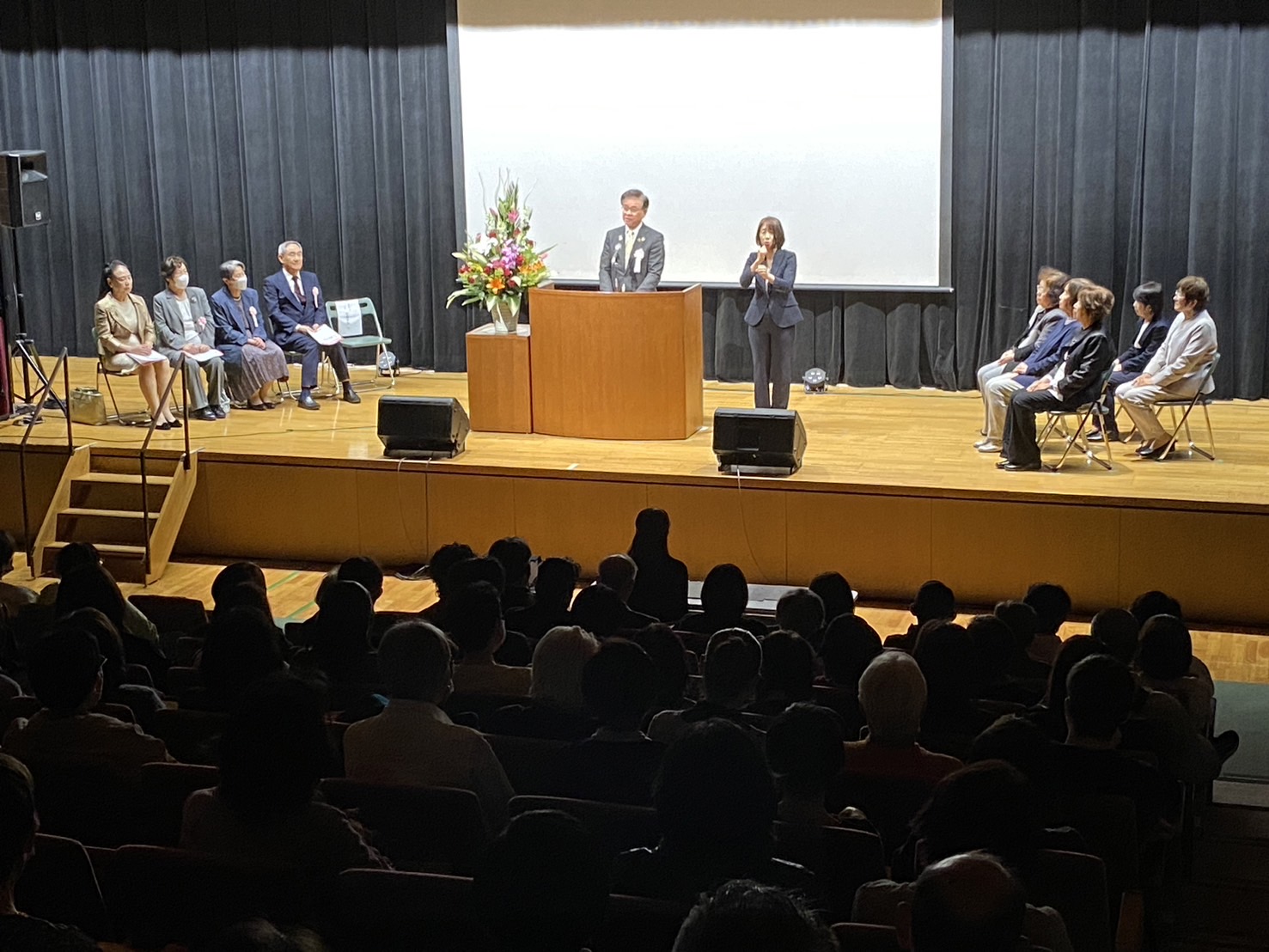 ステージ上の演台で、スーツ姿の市長がスピーチし横に立つ女性が話に合わせて手話をする様子や、両脇に来賓が、客席には大勢の聴衆が座っている式典全体の様子を写した写真