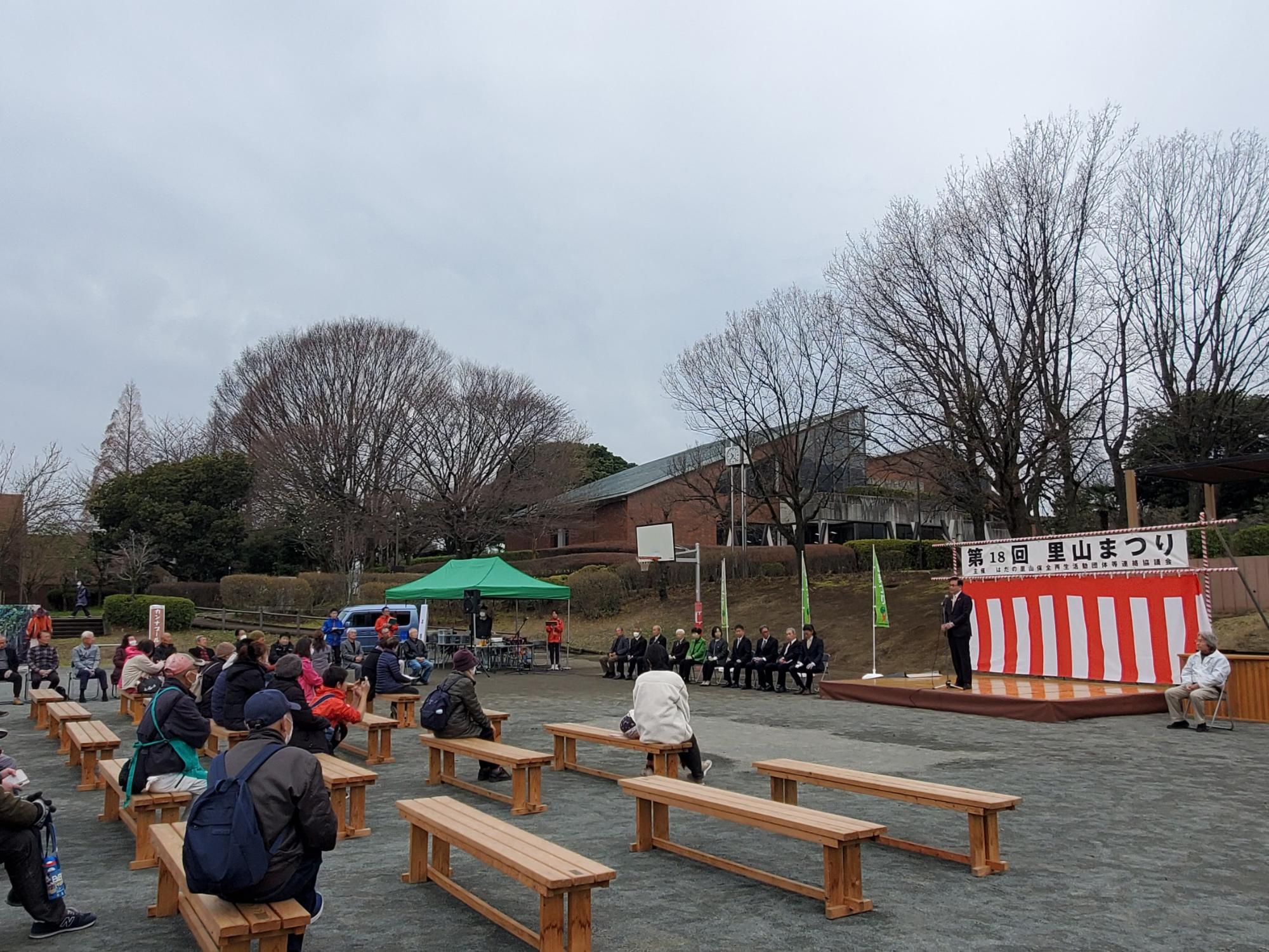 「里山まつり」のステージ上で市長が挨拶し、壇上横には来賓が、客席のベンチには多くの観客が座って聞いている様子を後方から写した写真
