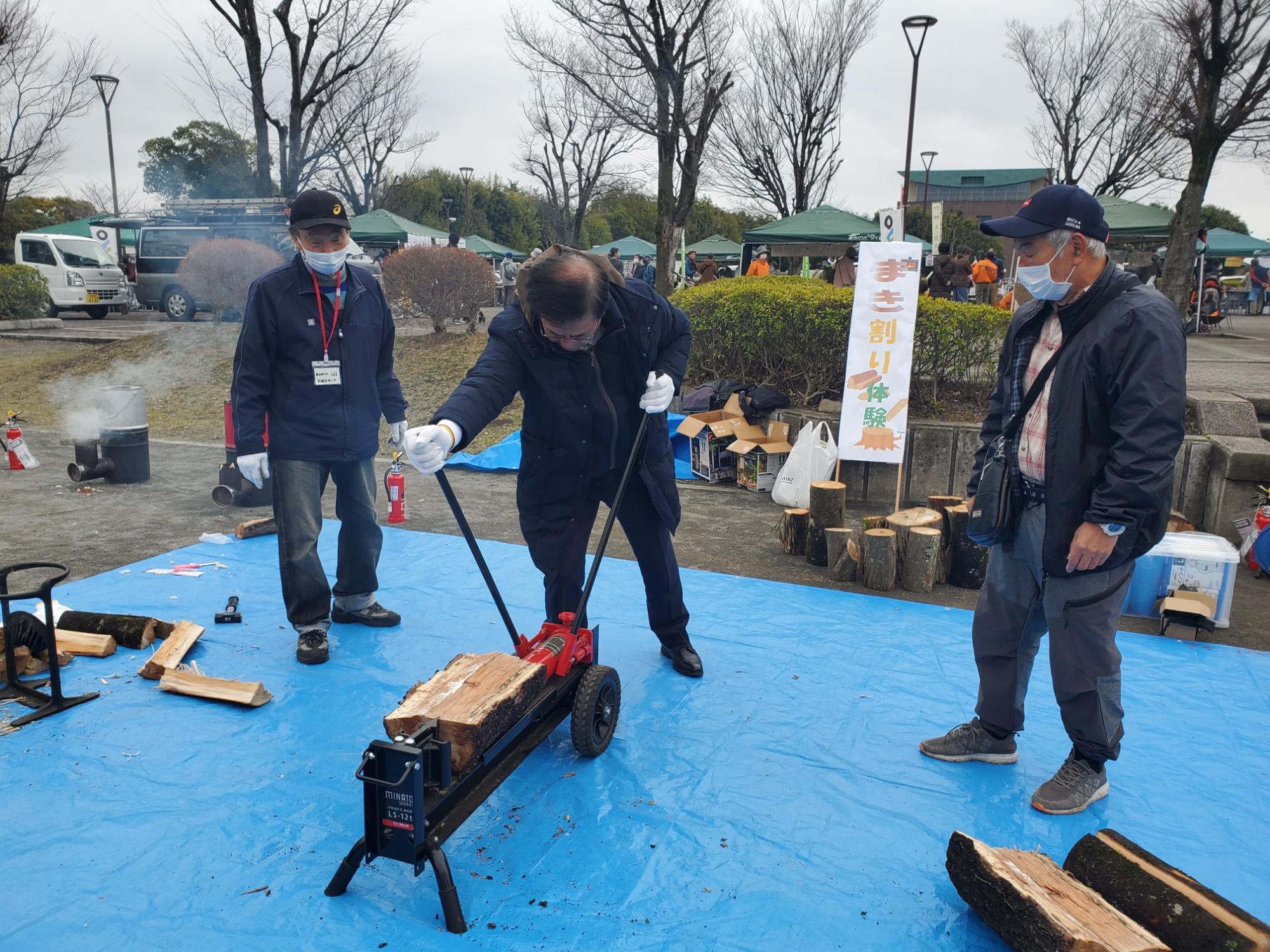 「薪割り体験」と書かれたブースのブルーシートの上で、市長が薪割り機を操作し、その様子を両脇の男性二人が見守っている写真