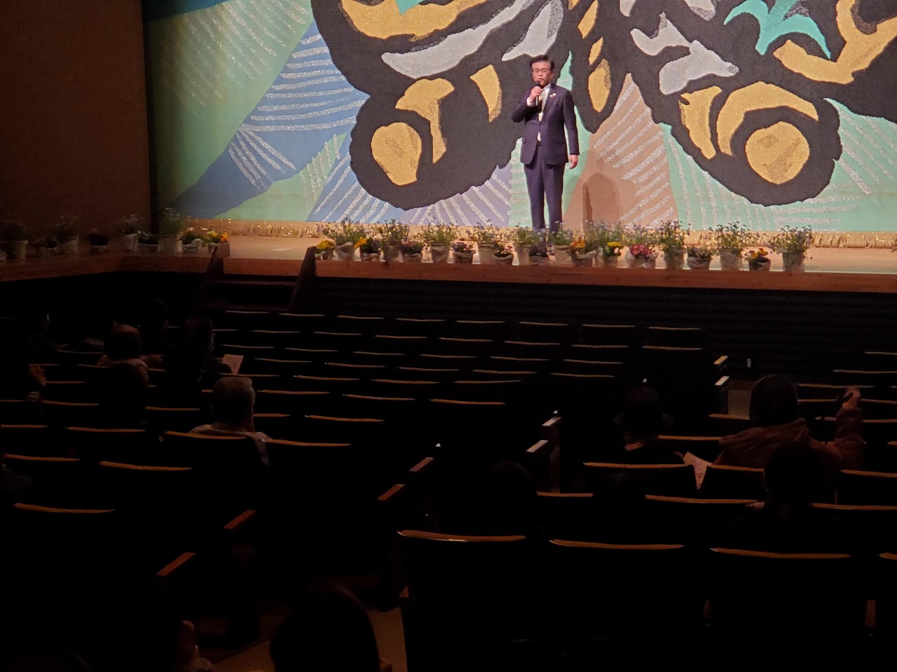 緞帳が降りた舞台上で、客席の方を向きスーツの市長が話をしている様子を後方から写した写真