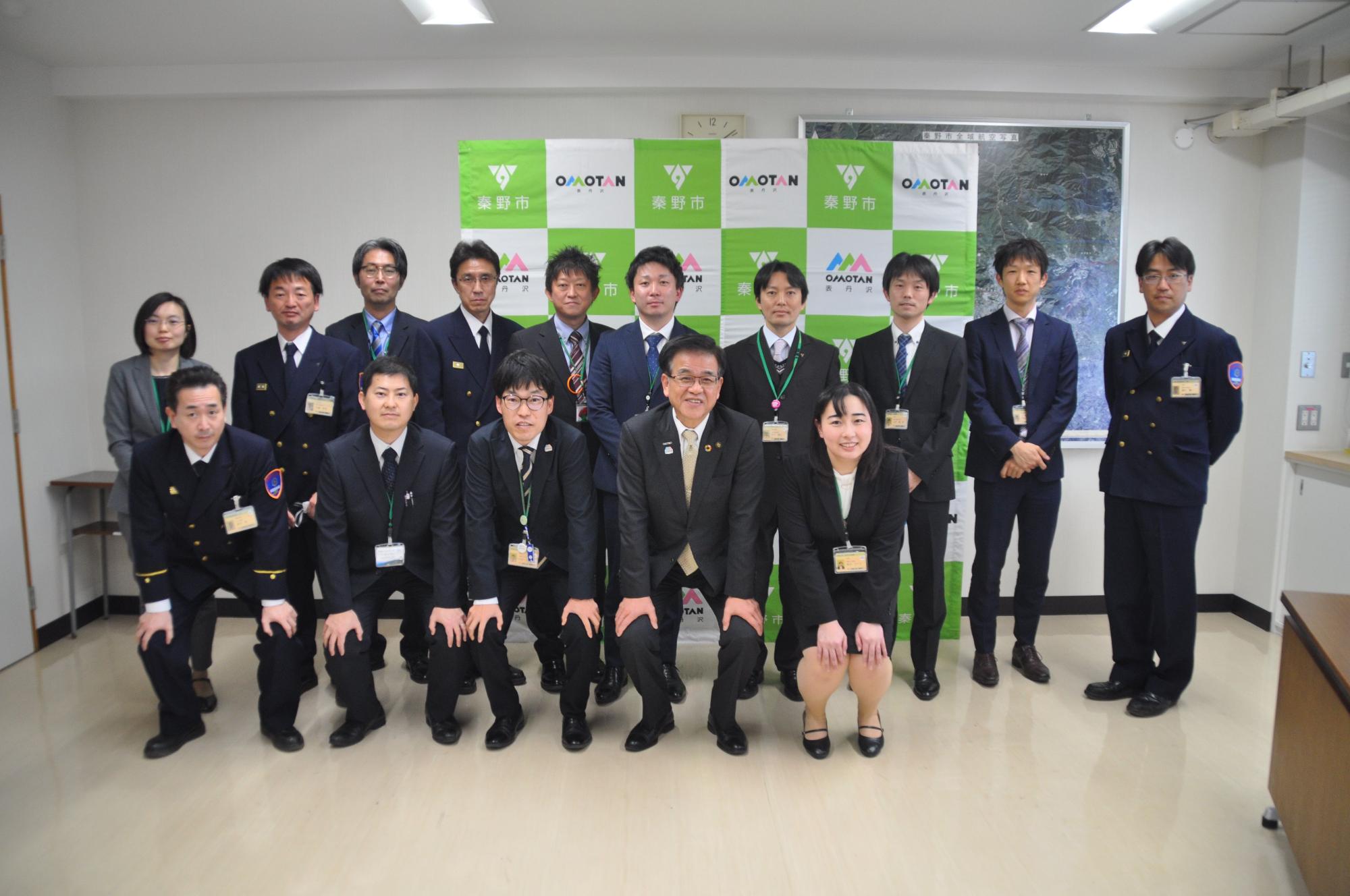 「秦野市」のパネルを背に、スーツや制服姿の職員の方々が、前列はしゃがみ、後列は立って記念撮影している写真