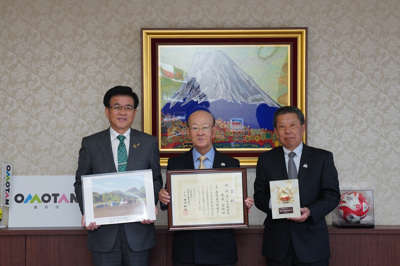 スーツの市長と栗原さんと関係者の方がそれぞれ写真、賞状、盾を手に持って横一列に並び、記念撮影している写真