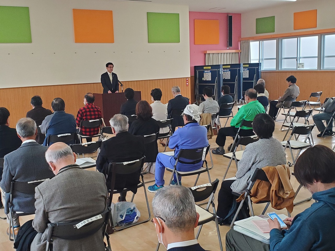 スーツ姿の市長が演台から話し、椅子に座った多くの地域住民と思われる人々が耳を傾けている写真