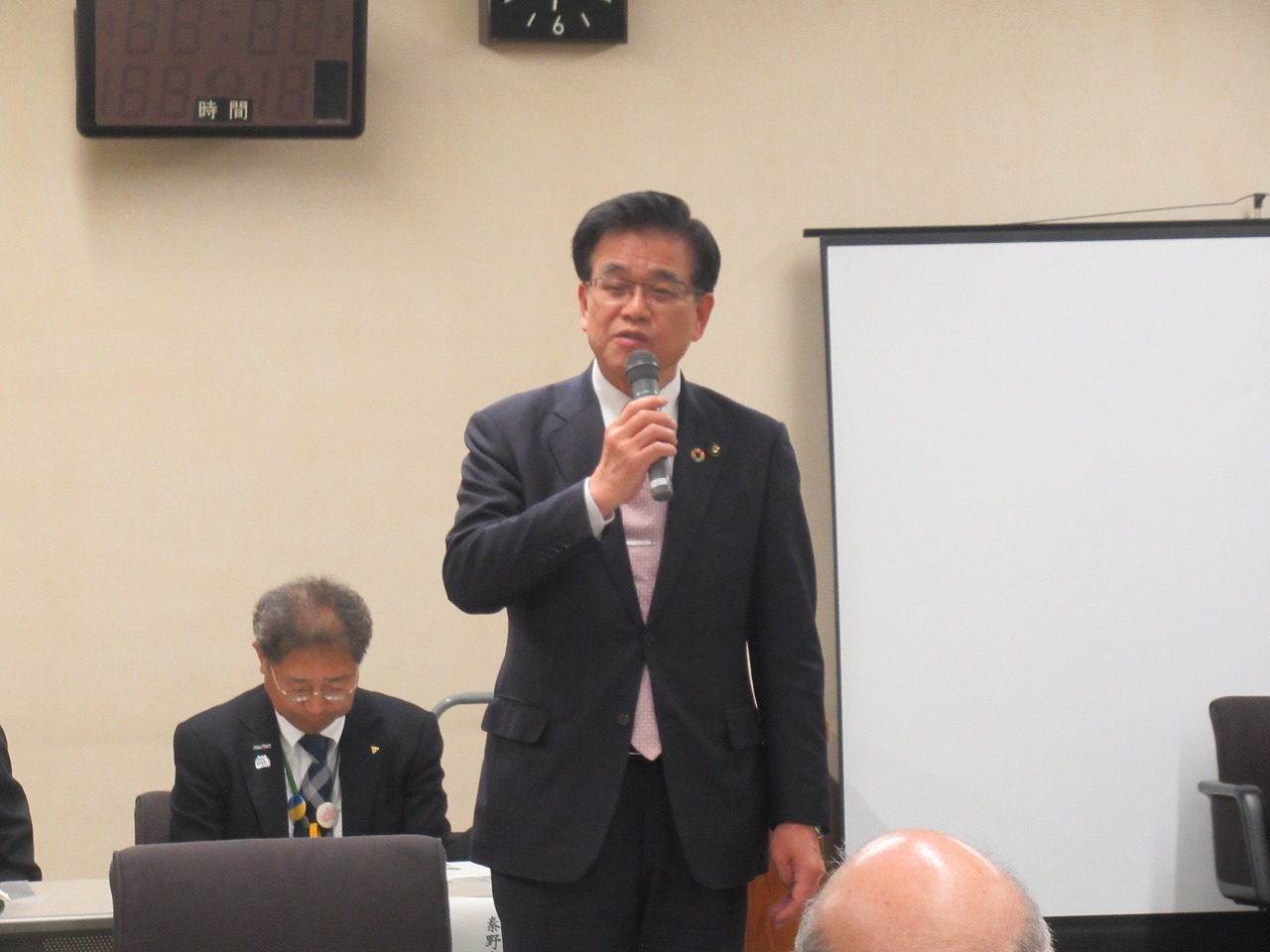 スクリーンが設置された会議室で、スーツ姿で眼鏡をかけた市長がマイクを手に持ち、真剣な表情で話をしている様子の写真