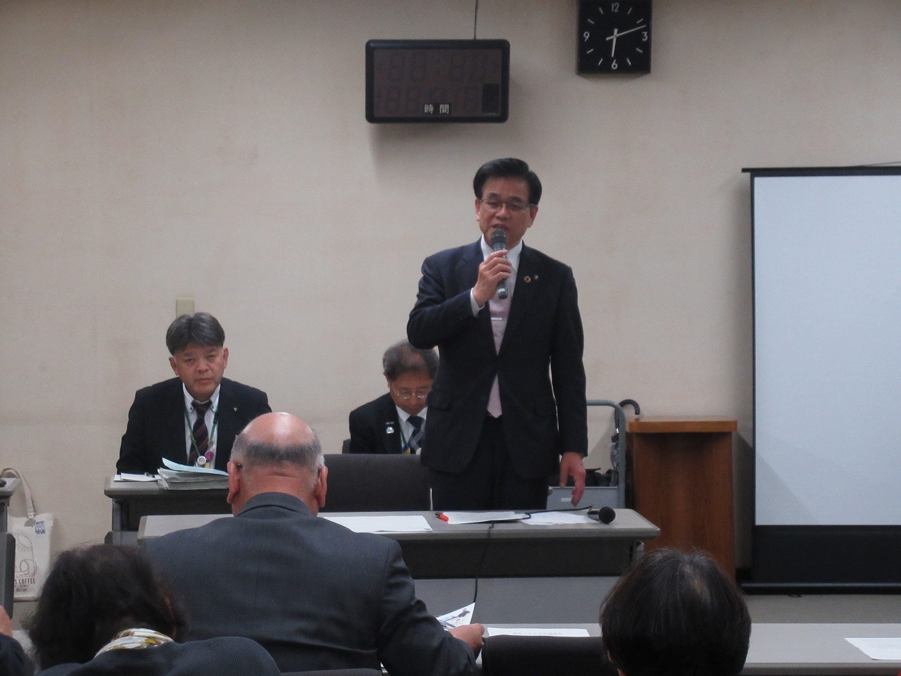 スーツ姿の市長が会議室でマイクを握って挨拶し、前方には設置された机に座り参加者が話を聞いている様子の写真