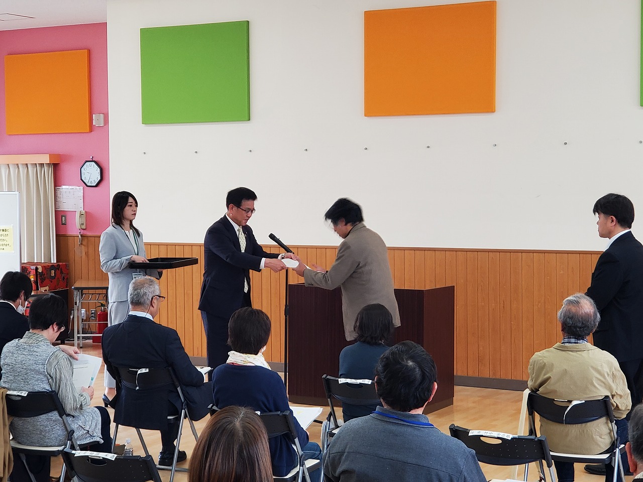 スーツ姿の市長からブラウン色のジャケットを着た男性へ委託状が手渡されている表彰式の様子を写した写真