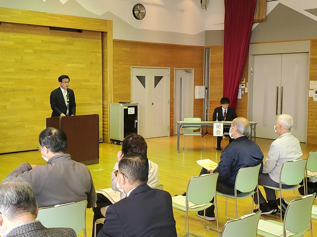 室内の前方に設置された演台でスーツ姿の市長がスピーチし、椅子に座った参加者が聞いている写真