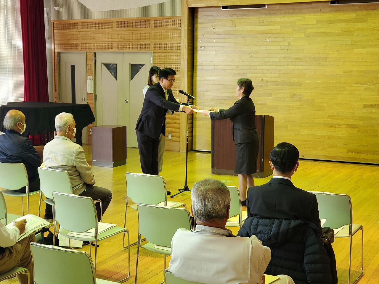 室内の前方に設置された演台の前でスーツ姿の市長が女性に委託状を手渡している写真