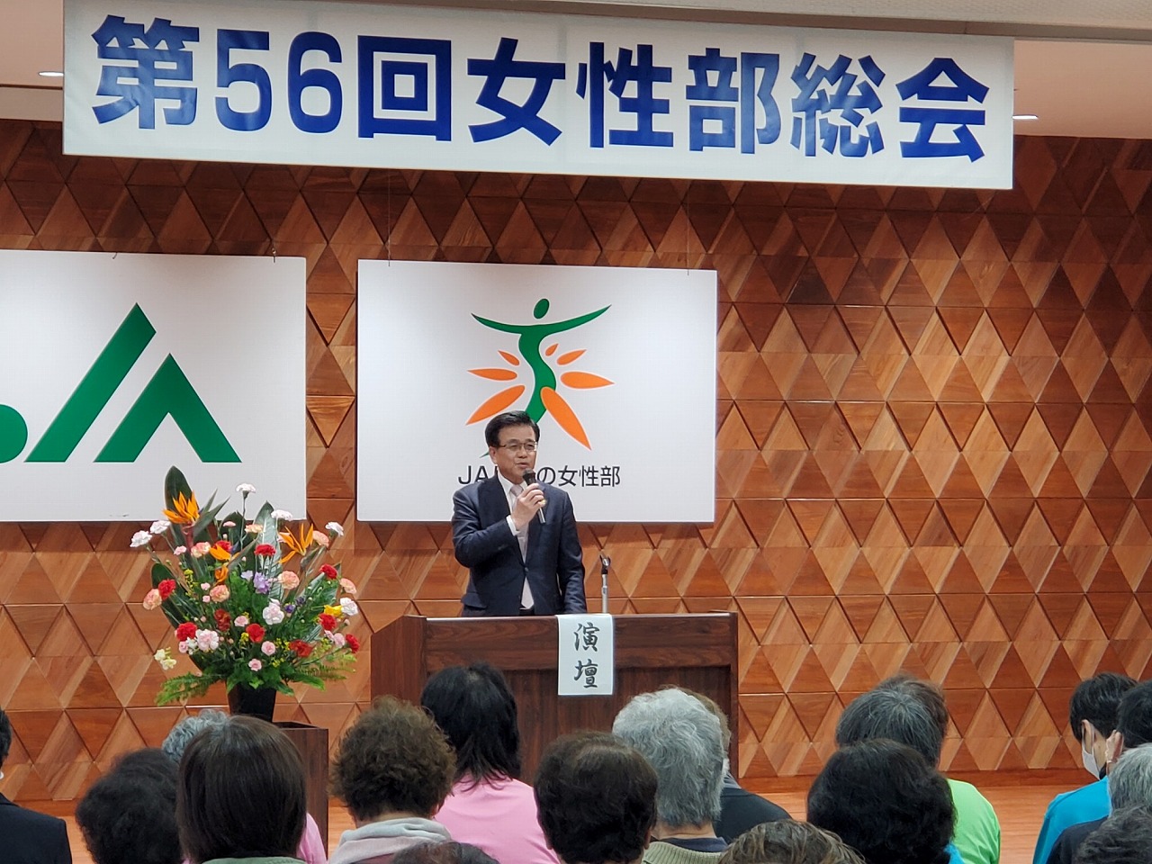 「第56回女性部総会」の横断幕が掲げられた会場の前方に設置された演台からスーツ姿の市長が参加者の方々に向かって話をしている写真