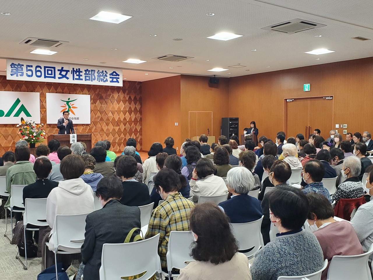 「女性部総会」の横断幕の下、スーツの市長が挨拶し、会場に設置された席に大勢の参加者が耳を傾けている様子を後方から写した写真